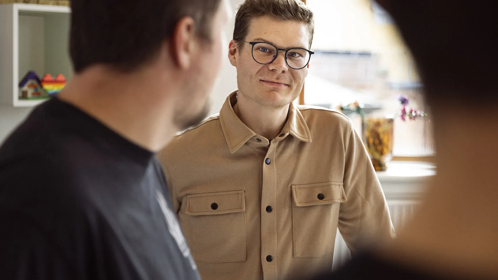 Billede af en ung mand med briller og et venligt smil, iført en beige skjorte med lommer, der står i et lyst og indbydende rum, mens han interagerer med andre personer i en uformel samtale.