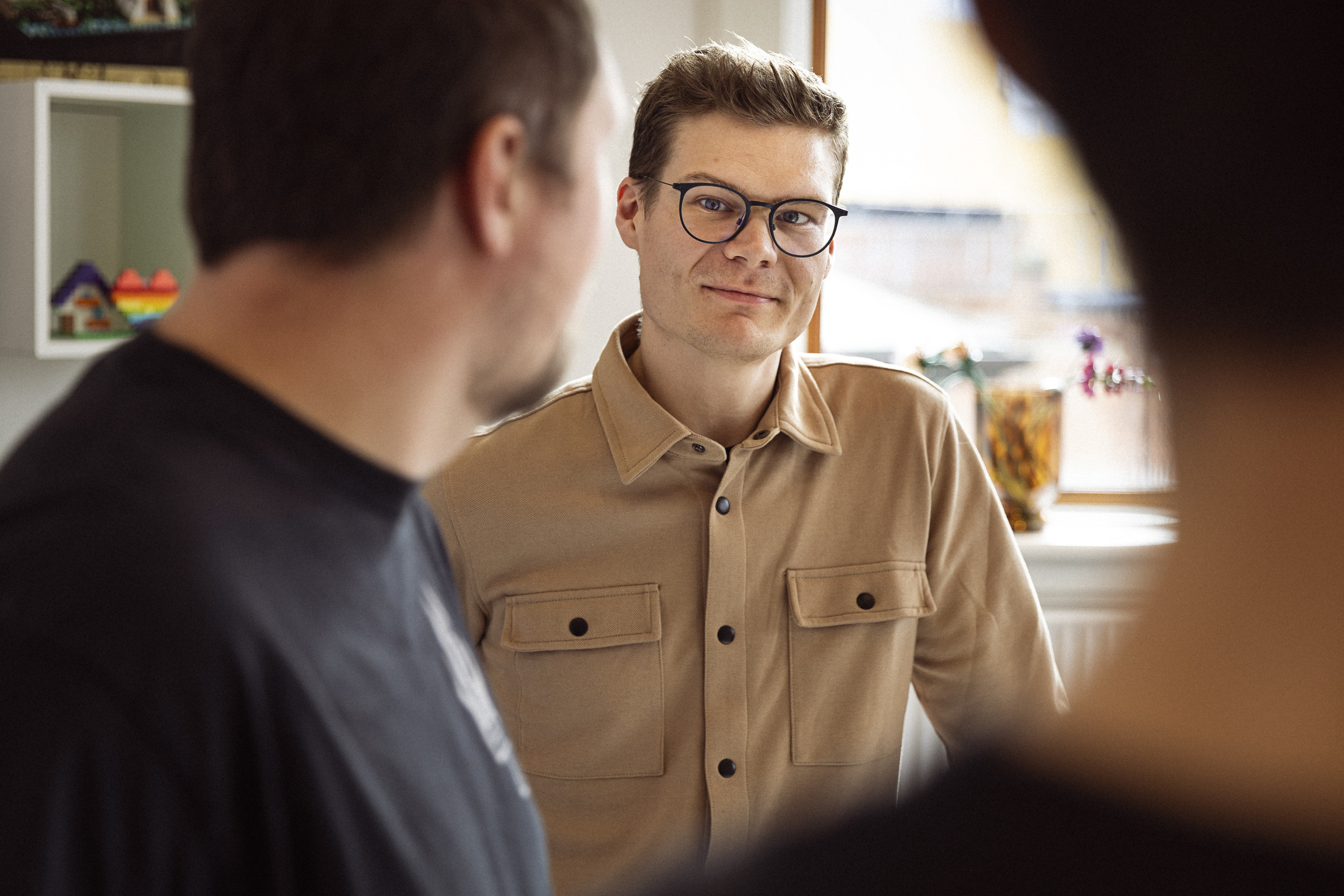 Billede af en ung mand med briller og et venligt smil, iført en beige skjorte med lommer, der står i et lyst og indbydende rum, mens han interagerer med andre personer i en uformel samtale.