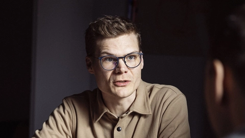 A man with glasses and a beige shirt engaged in a conversation at a table surrounded by smartphones and notes, highlighting a collaborative work environment.