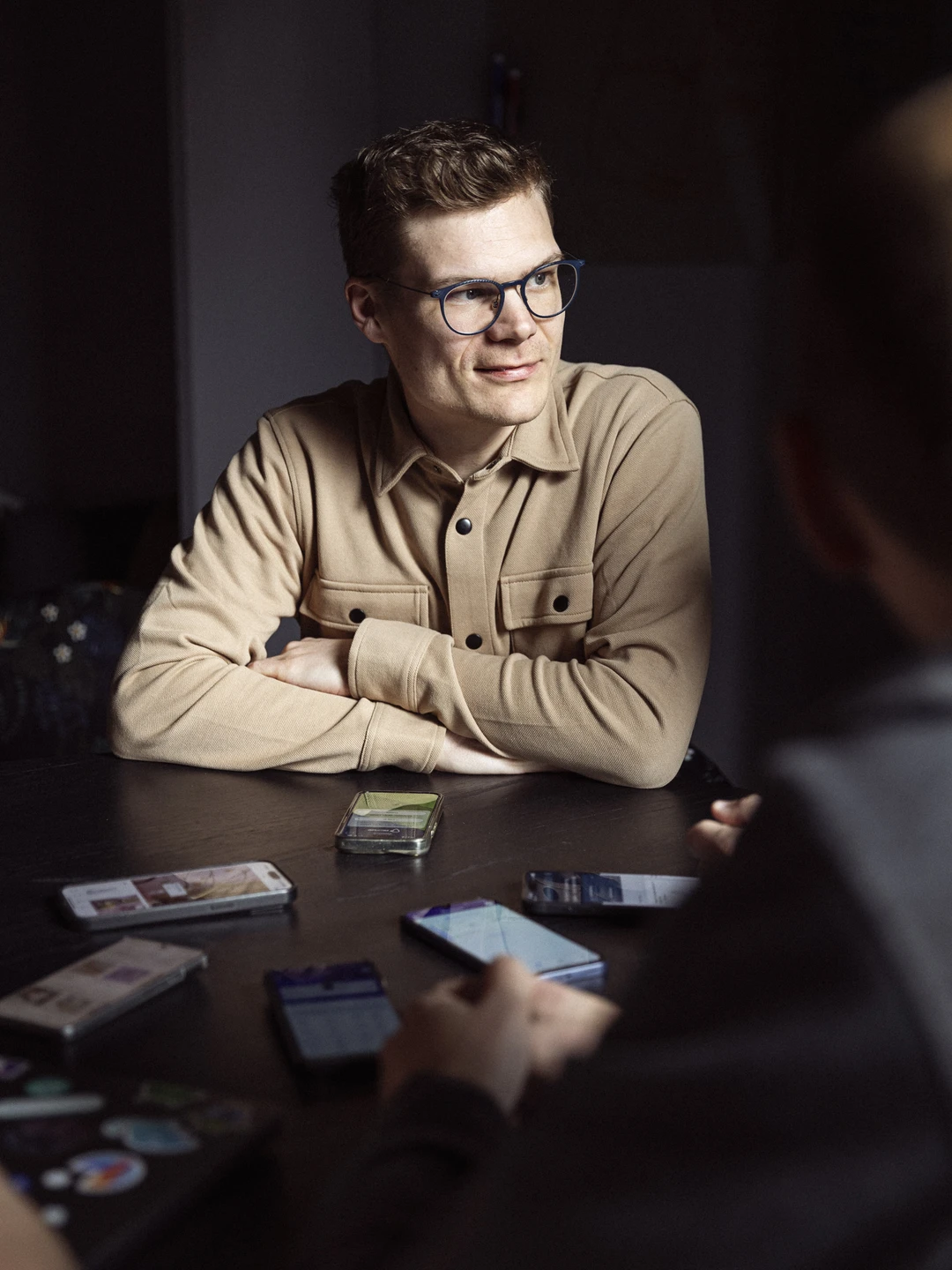 Ung mand med brune krøller og briller, iført en beige skjorte, sidder ved et bord med flere smartphones, mens han smiler og kigger op.
