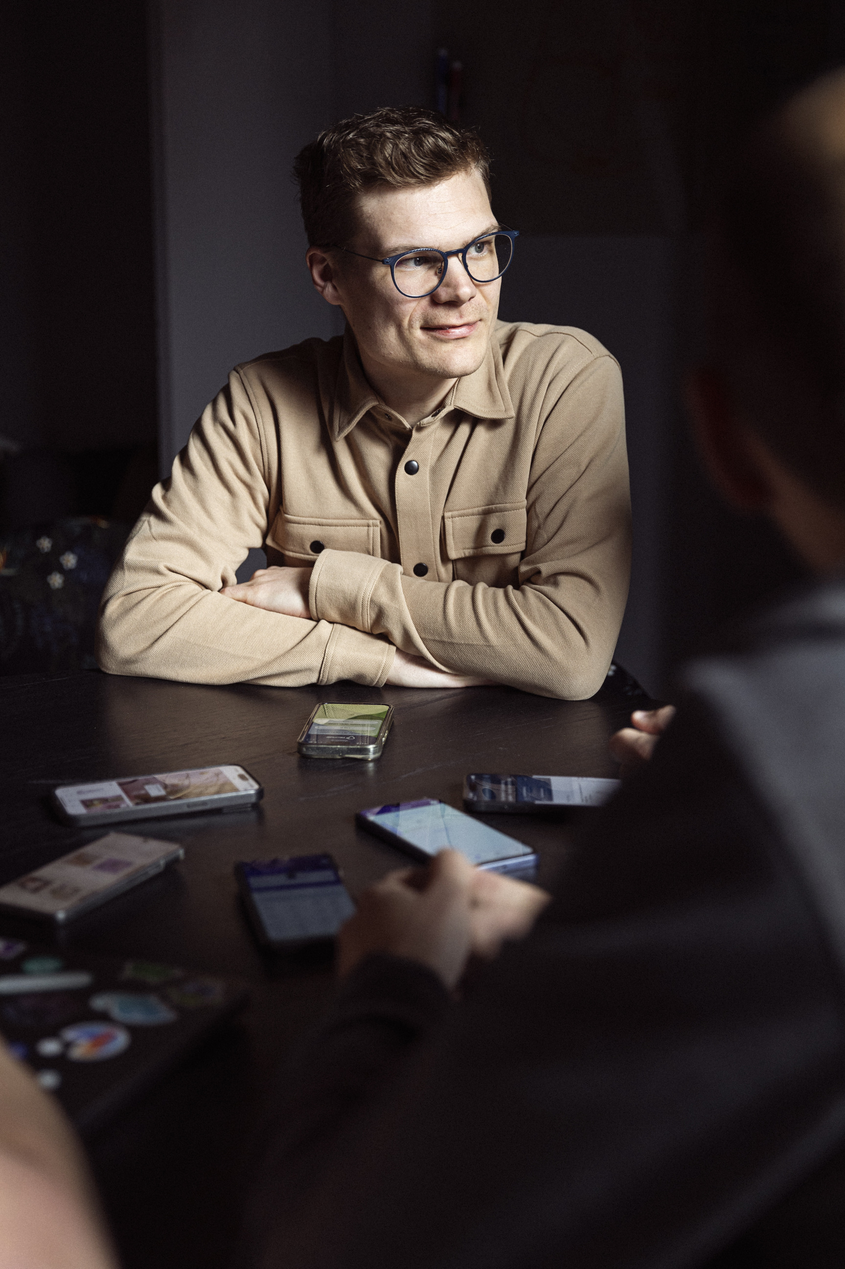 Ung mand med brune krøller og briller, iført en beige skjorte, sidder ved et bord med flere smartphones, mens han smiler og kigger op.