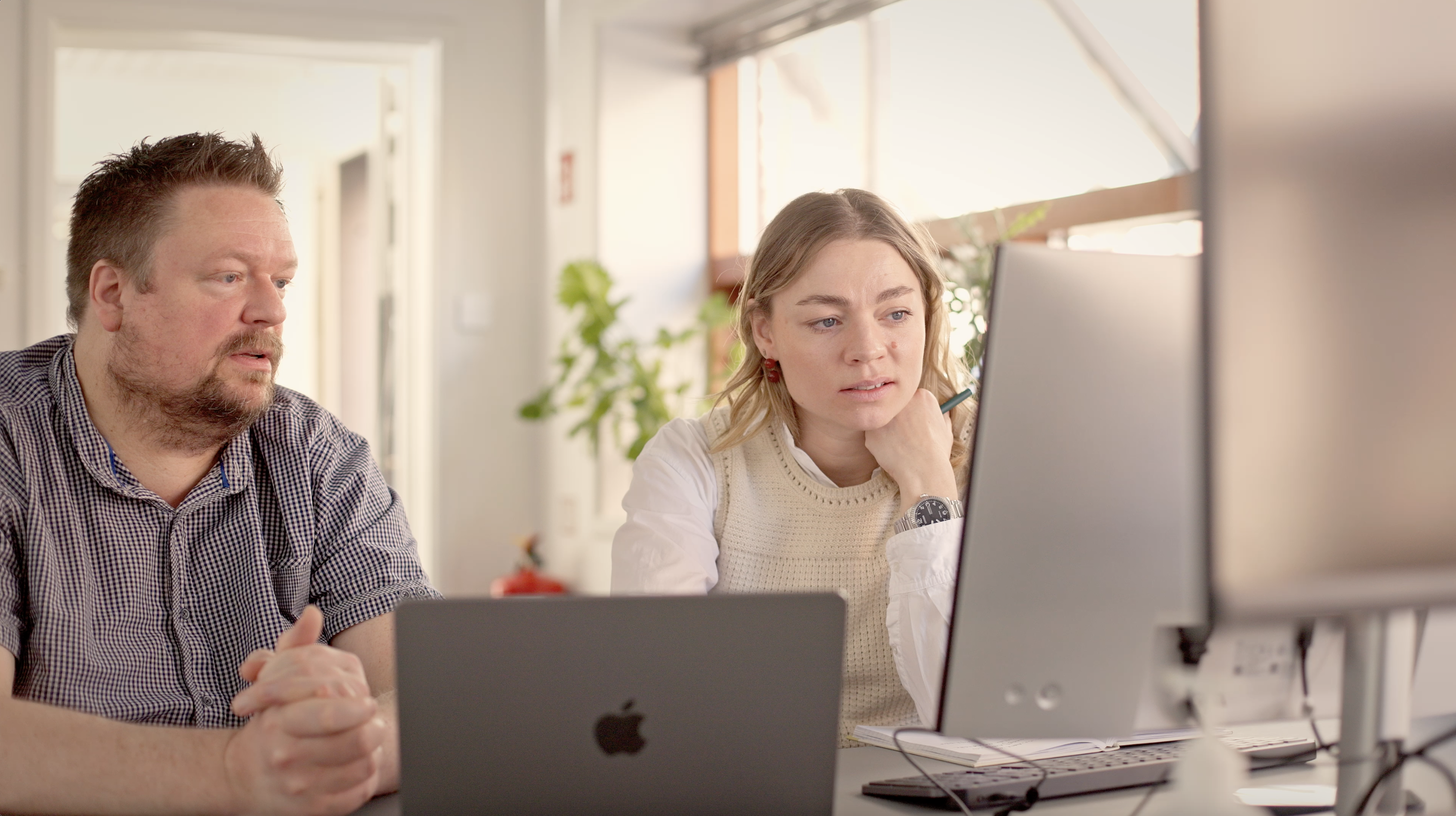 To personer sidder ved et bord med computere, en mand med kort hår og skæg, en kvinde med bølget hår, begge fokuseret på skærmen, der er planter i baggrunden og et moderne kontormiljø.