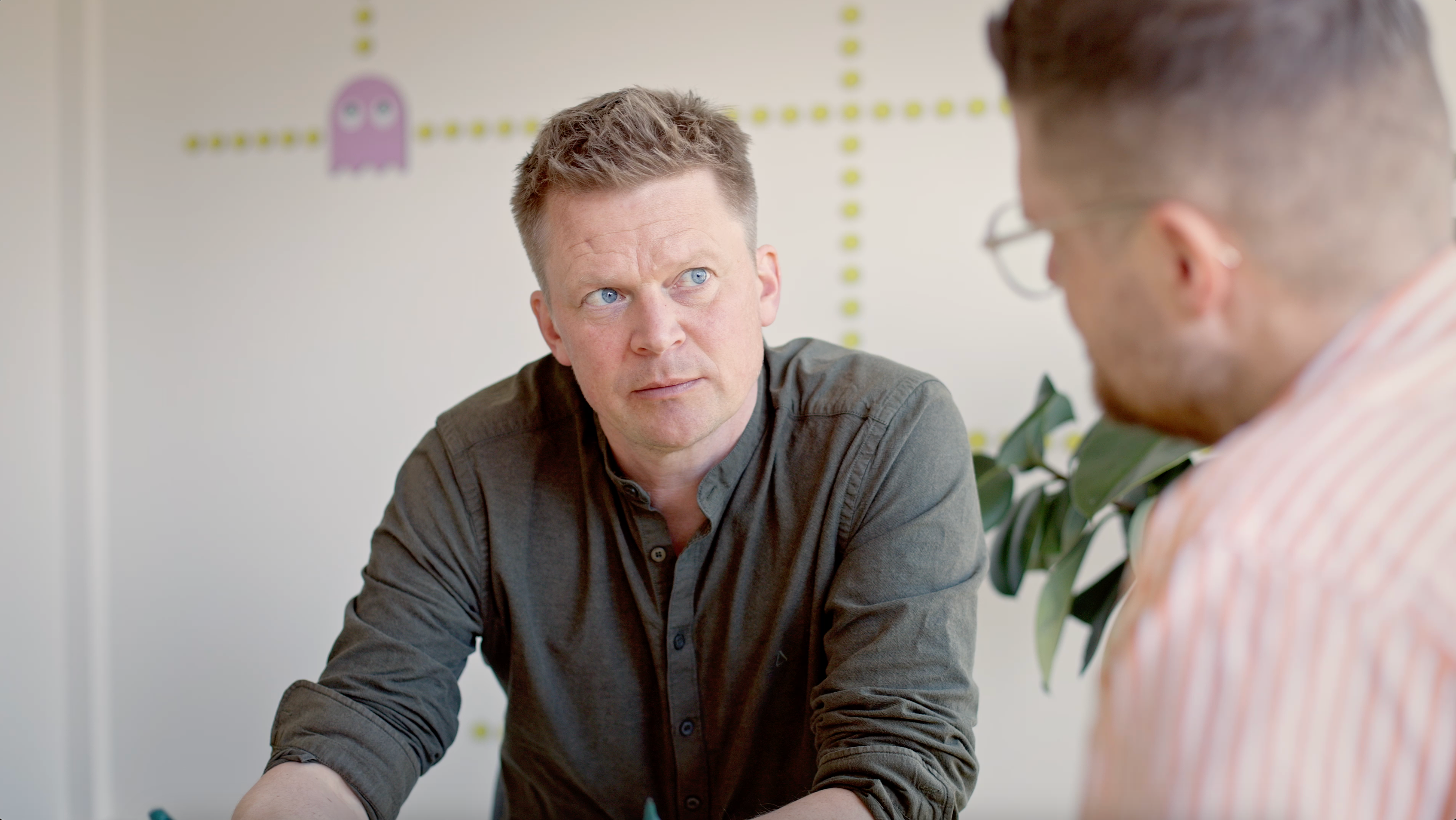 To mænd i en moderne kontorindstilling, den ene ser eftertænksom ud i en grøn skjorte, mens den anden, med briller og en stribet skjorte, koncentrerer sig om samtalen, med planter og kunst på væggen i baggrunden.