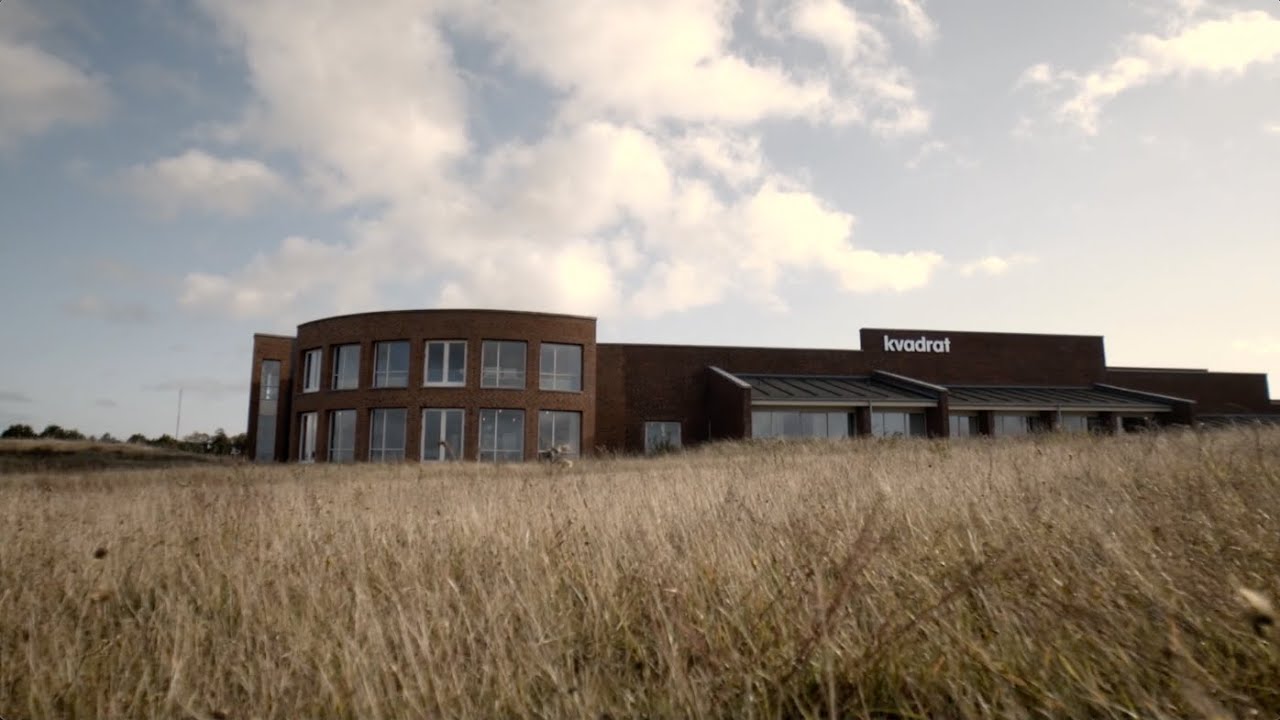 Billede af Kvadrat, en moderne bygning i et åbent landskab med højt græs og en skyet himmel. Bygningen har store vinduer og en karakteristisk, buet facade.