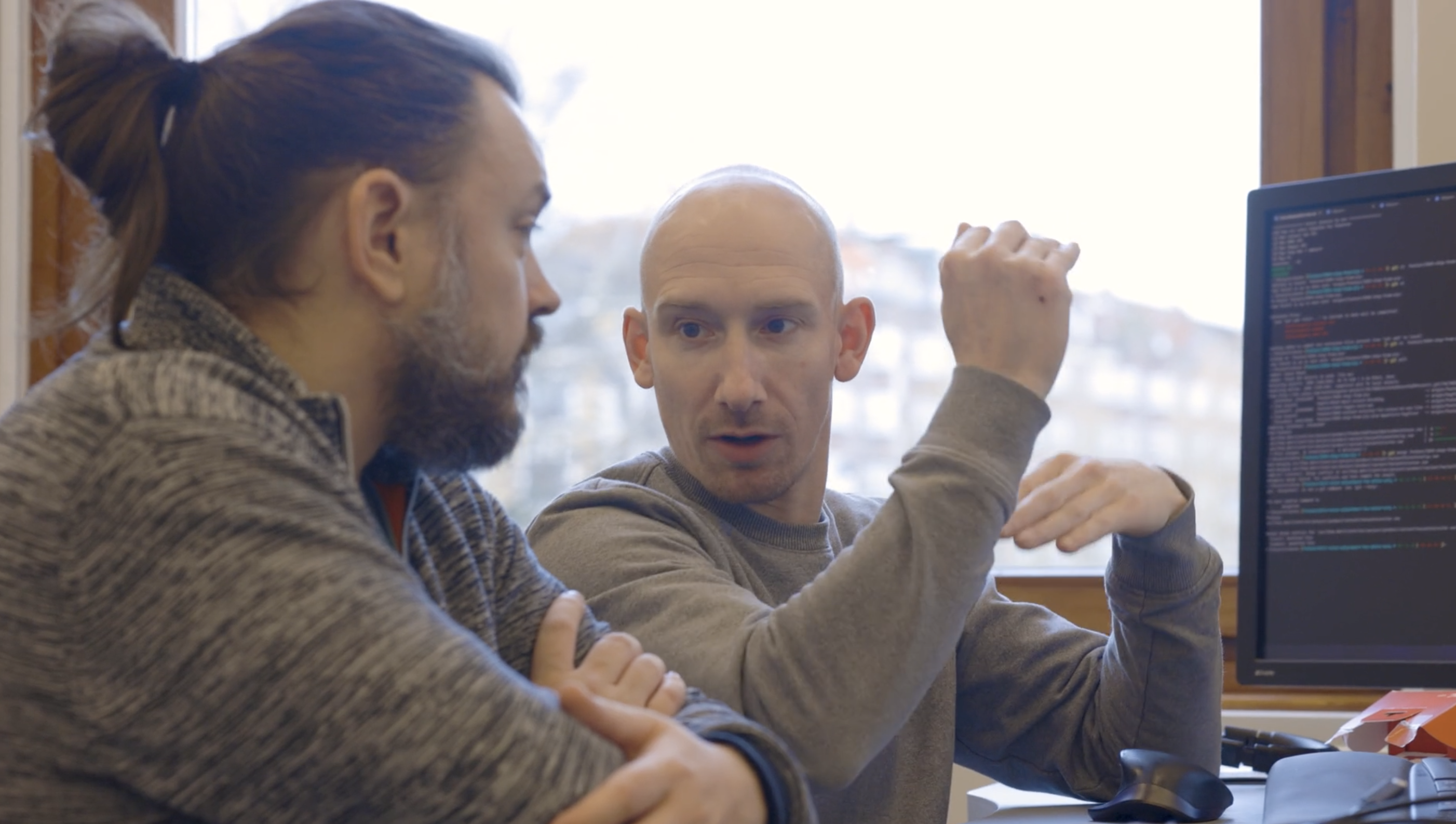 To mænd diskuterer engageret ved et skrivebord, mens de arbejder på en computer med kode på skærmen.