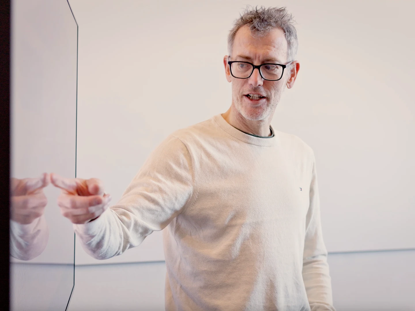 En mand i en lys sweater står ved siden af en skærm og peger med sin hånd, mens han forklarer noget.