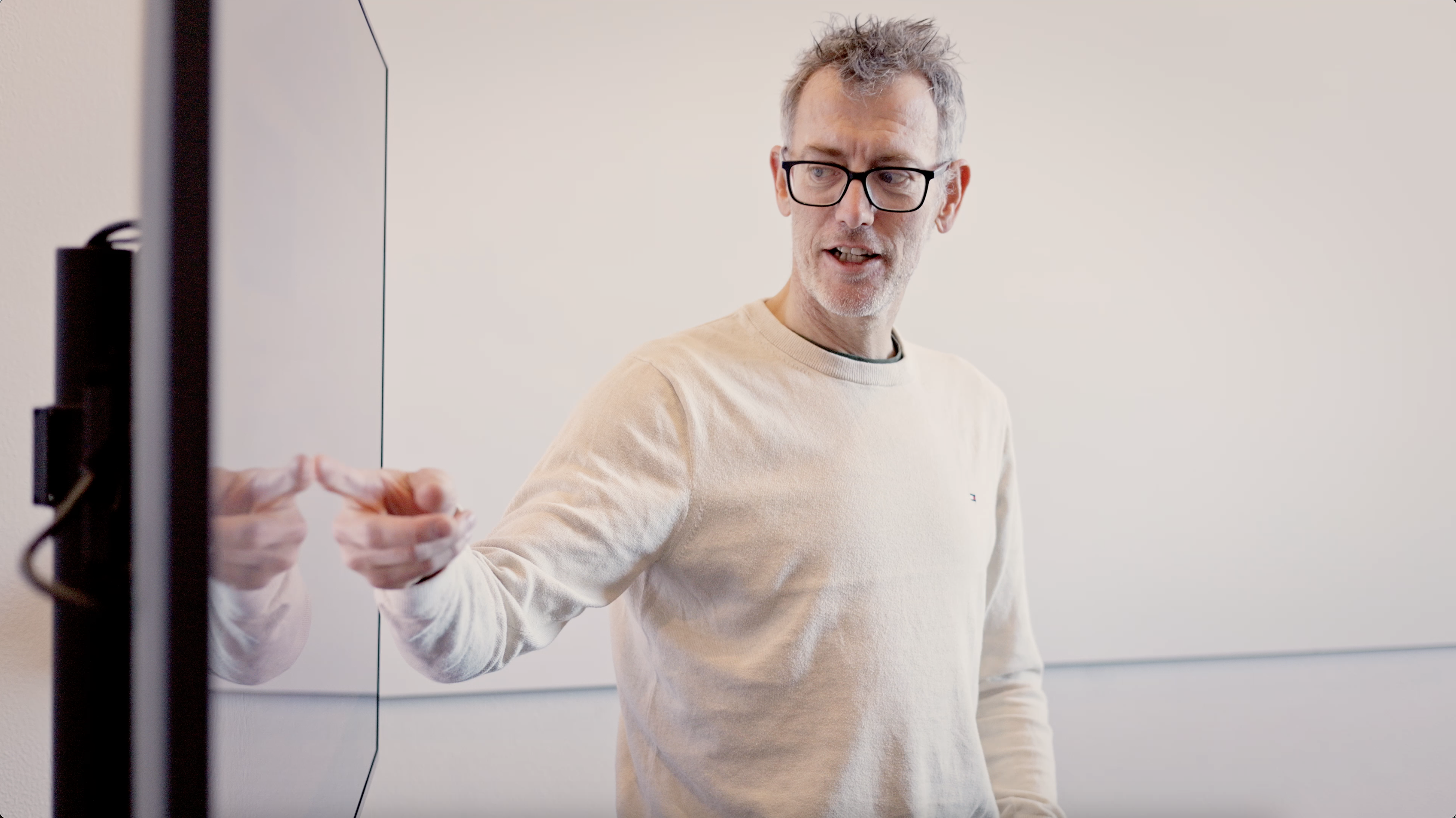 En mand i en lys sweater står ved siden af en skærm og peger med sin hånd, mens han forklarer noget.