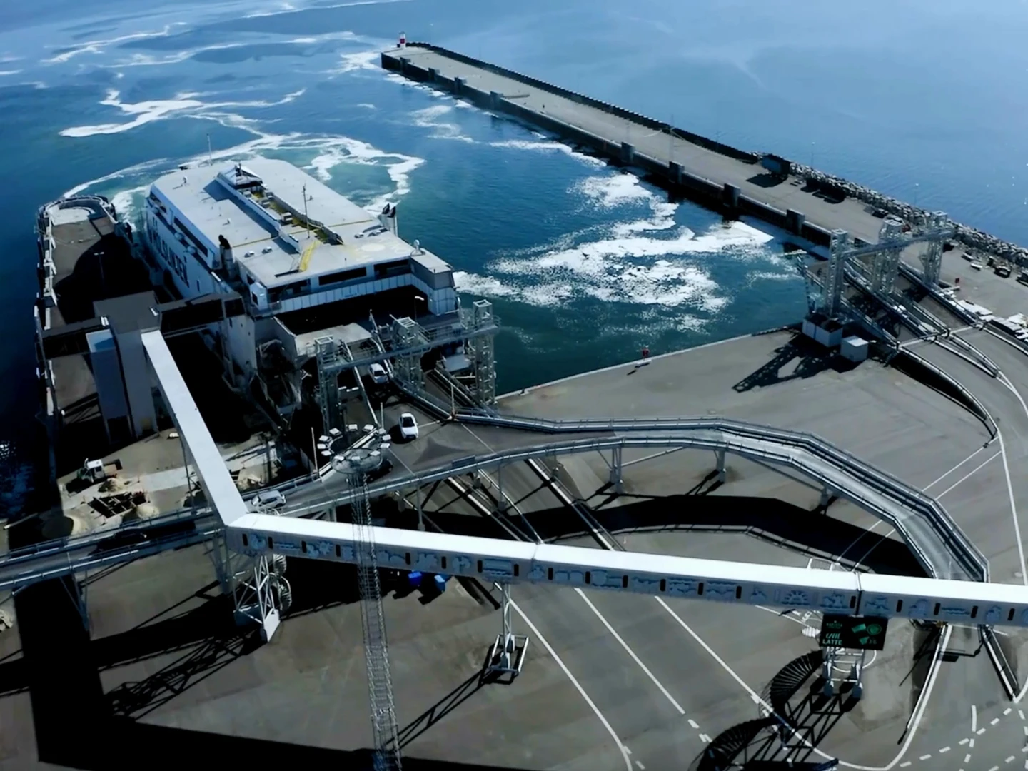Aerial view of a busy dock with ferries, cargo loading equipment, and a long pier extending into the ocean, showcasing industrial maritime operations and the surrounding water.