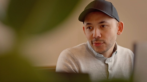 A focused man in a gray sweater and black cap is engaged in conversation, with green plant leaves slightly blurred in the foreground.