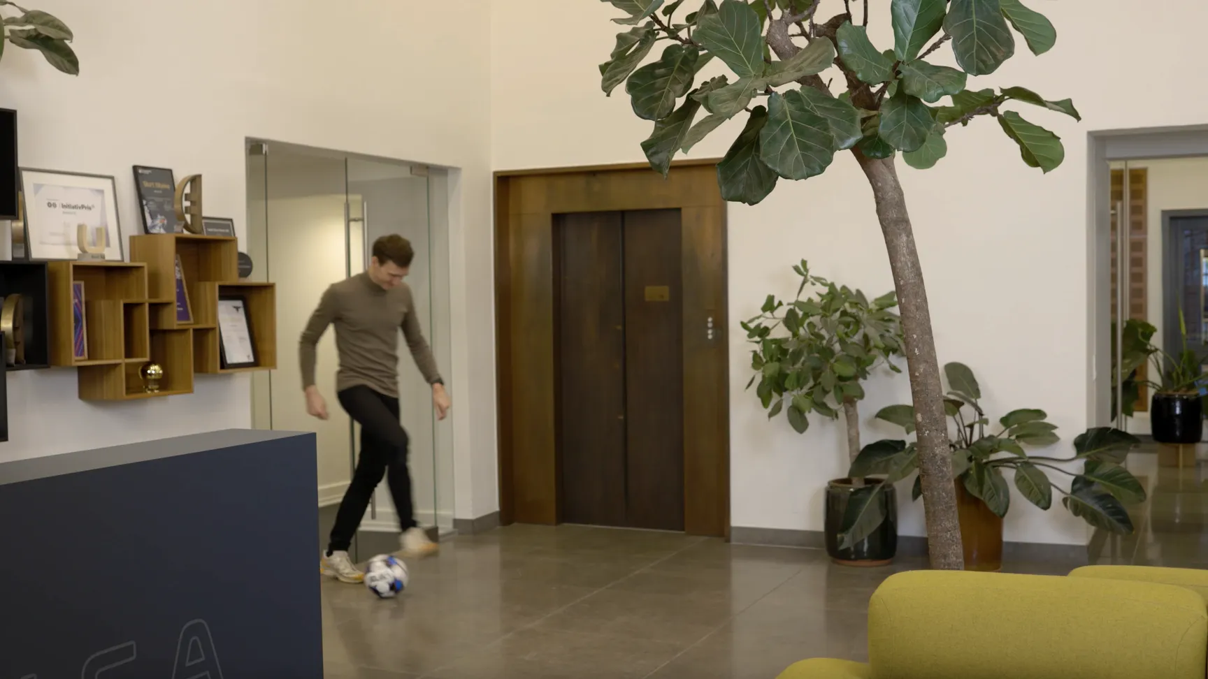 Mand spiller fodbold i en moderne kontorindretning med planter og et elevatorområde.