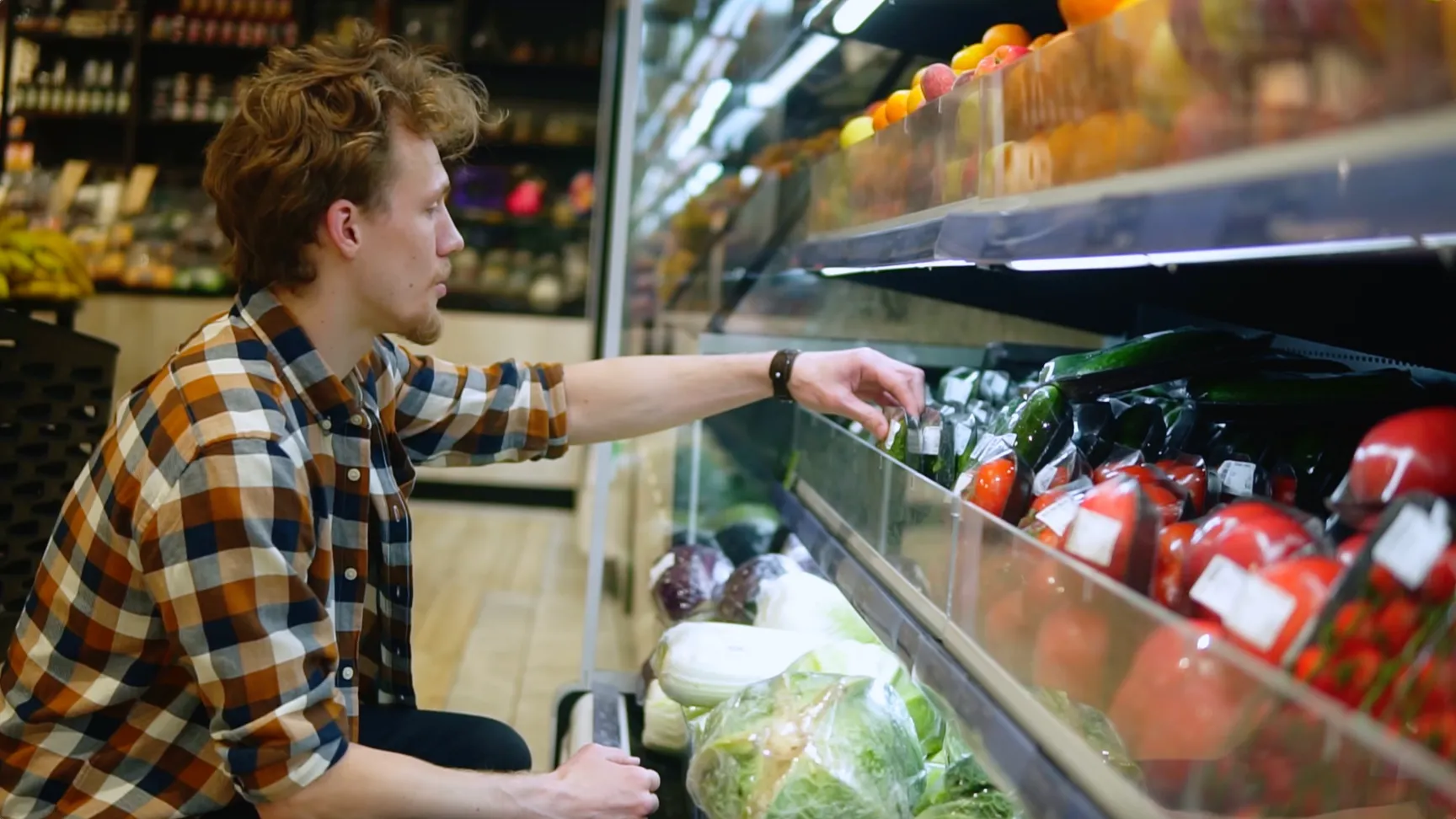 Young man in a plaid shirt selecting fresh vegetables in a grocery store, surrounded by various fruits and packaged produce.