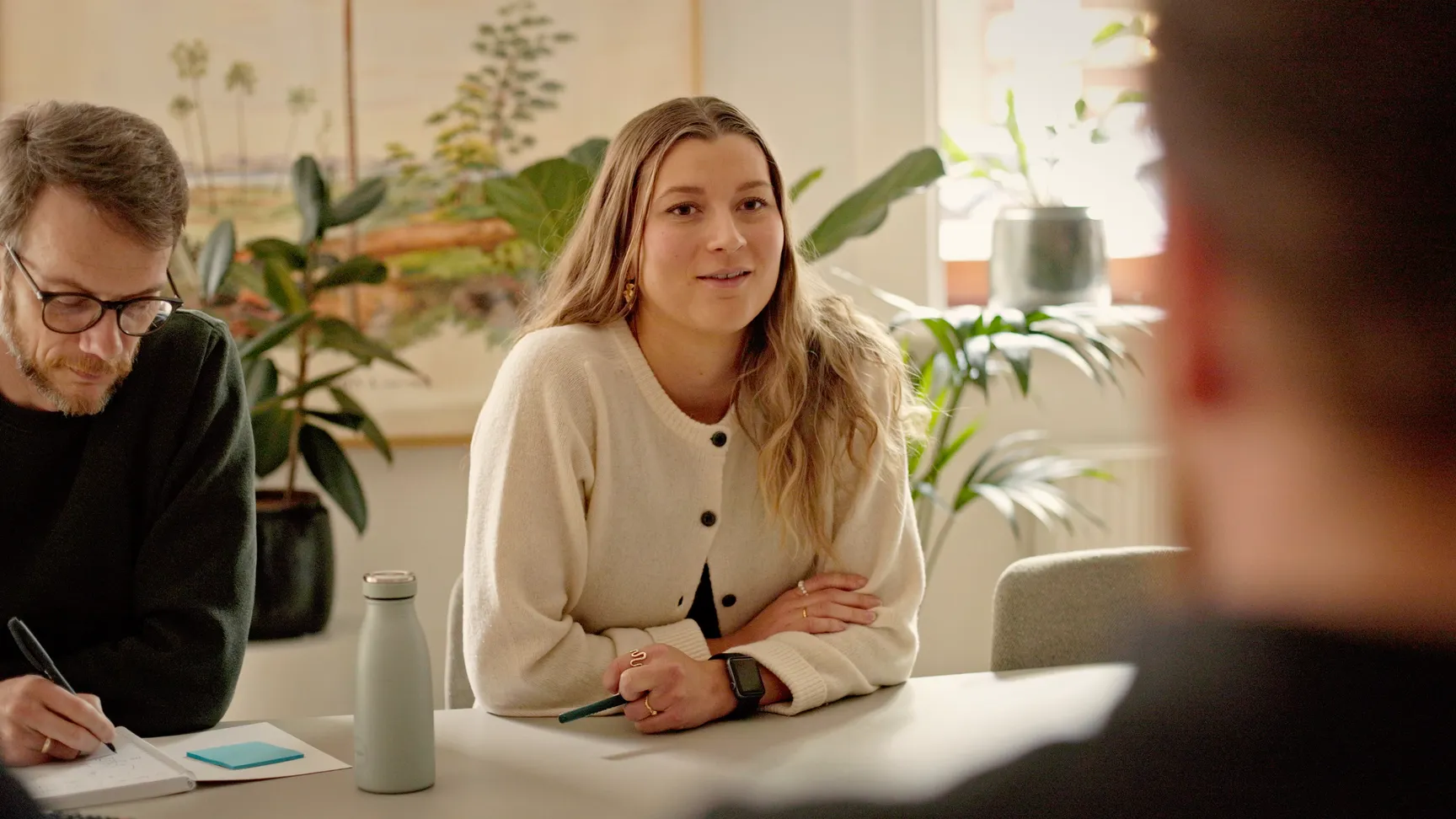 Ung kvinde med langt, bølget hår, iført en lys cardigan, deltager i et møde ved et bord. Hun smiler og ser op, mens en mand ved siden af skriver noter. Planter er synlige i baggrunden, som skaber en hyggelig atmosfære.