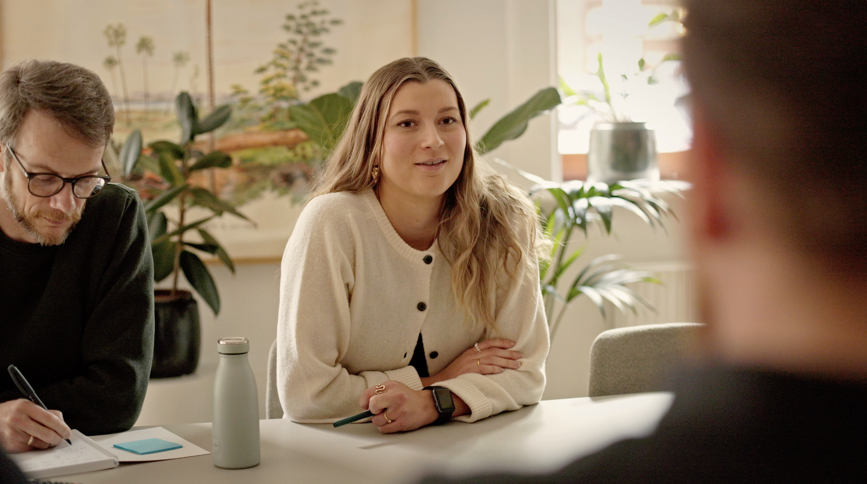 Ung kvinde med langt, bølget hår, iført en lys cardigan, deltager i et møde ved et bord. Hun smiler og ser op, mens en mand ved siden af skriver noter. Planter er synlige i baggrunden, som skaber en hyggelig atmosfære.