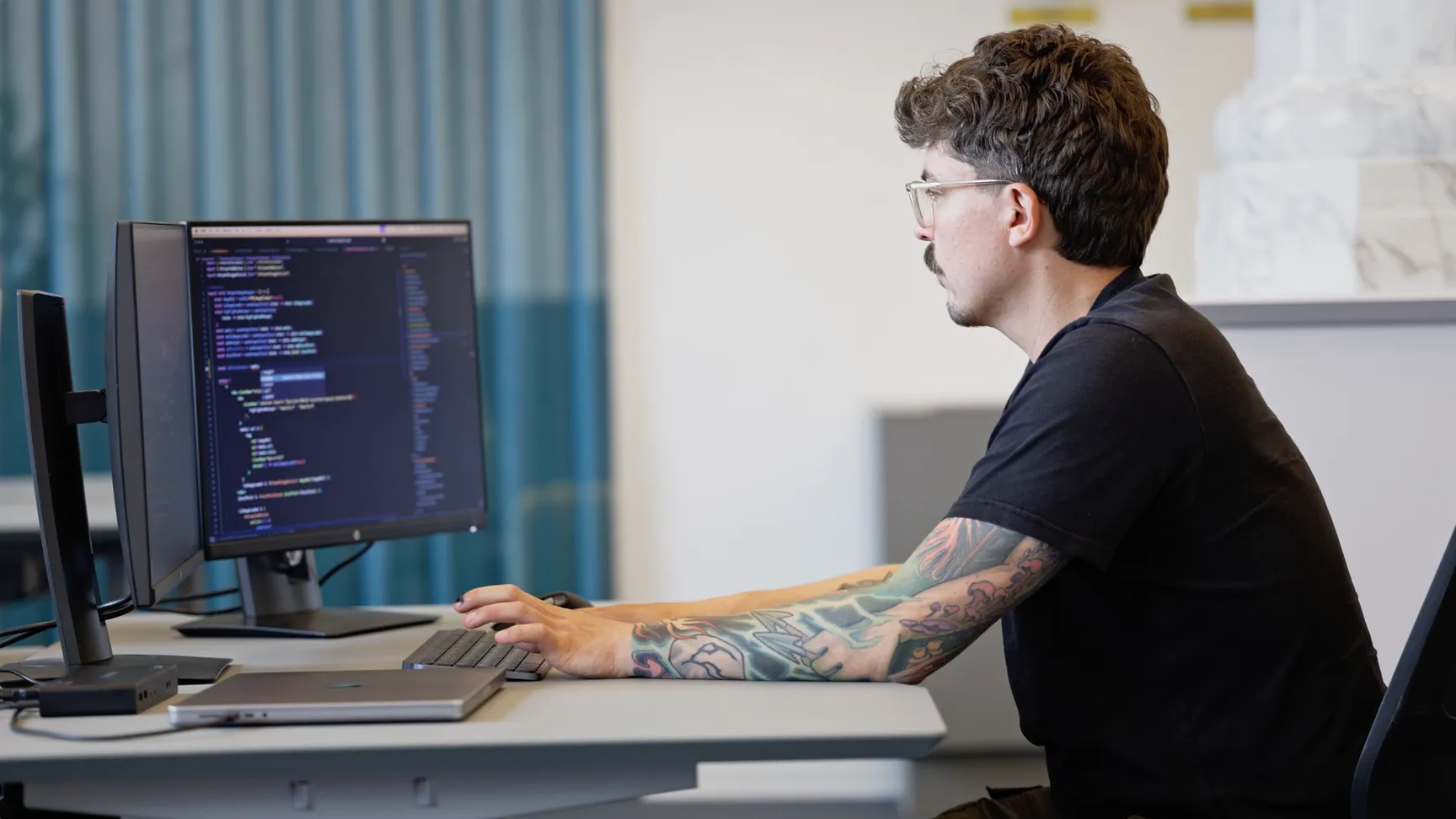 En programmør med tatoveringer sidder ved et skrivebord og arbejder på en computer med kode vist på skærmen.