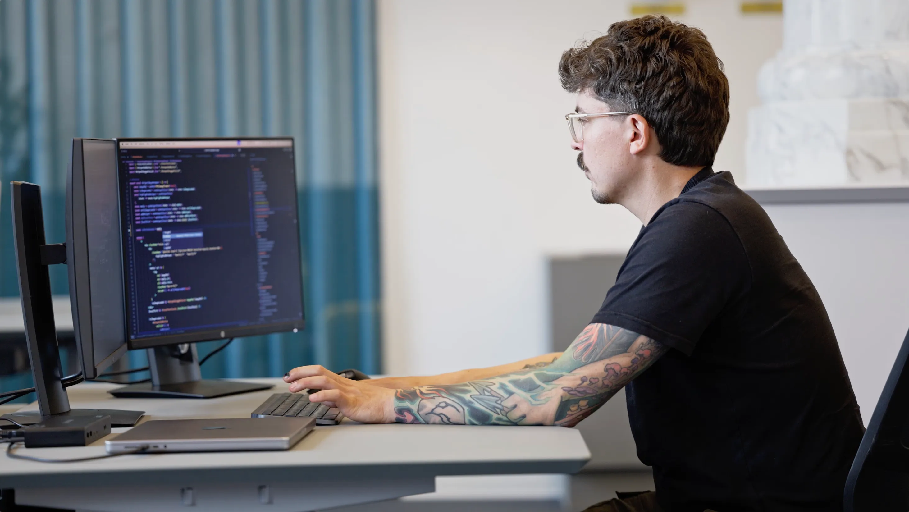 En programmør med tatoveringer sidder ved et skrivebord og arbejder på en computer med kode vist på skærmen.