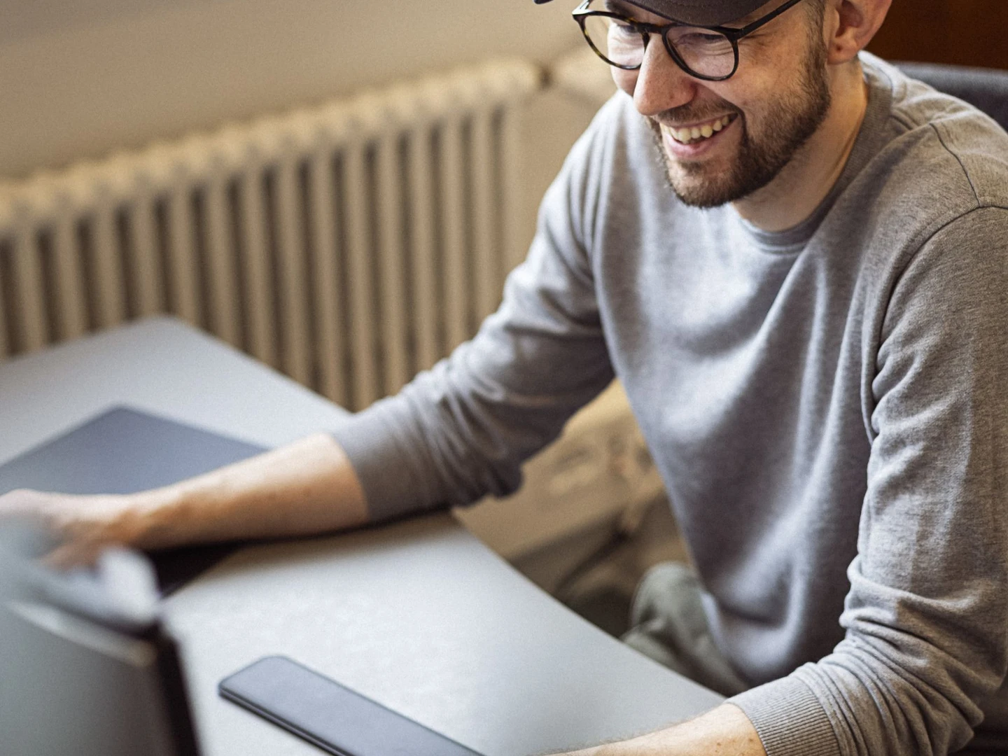 En mand med briller og kasket sidder foran en computer, smilende, mens han arbejder på en kontorarbejdsplads med en smartphone ved siden af.