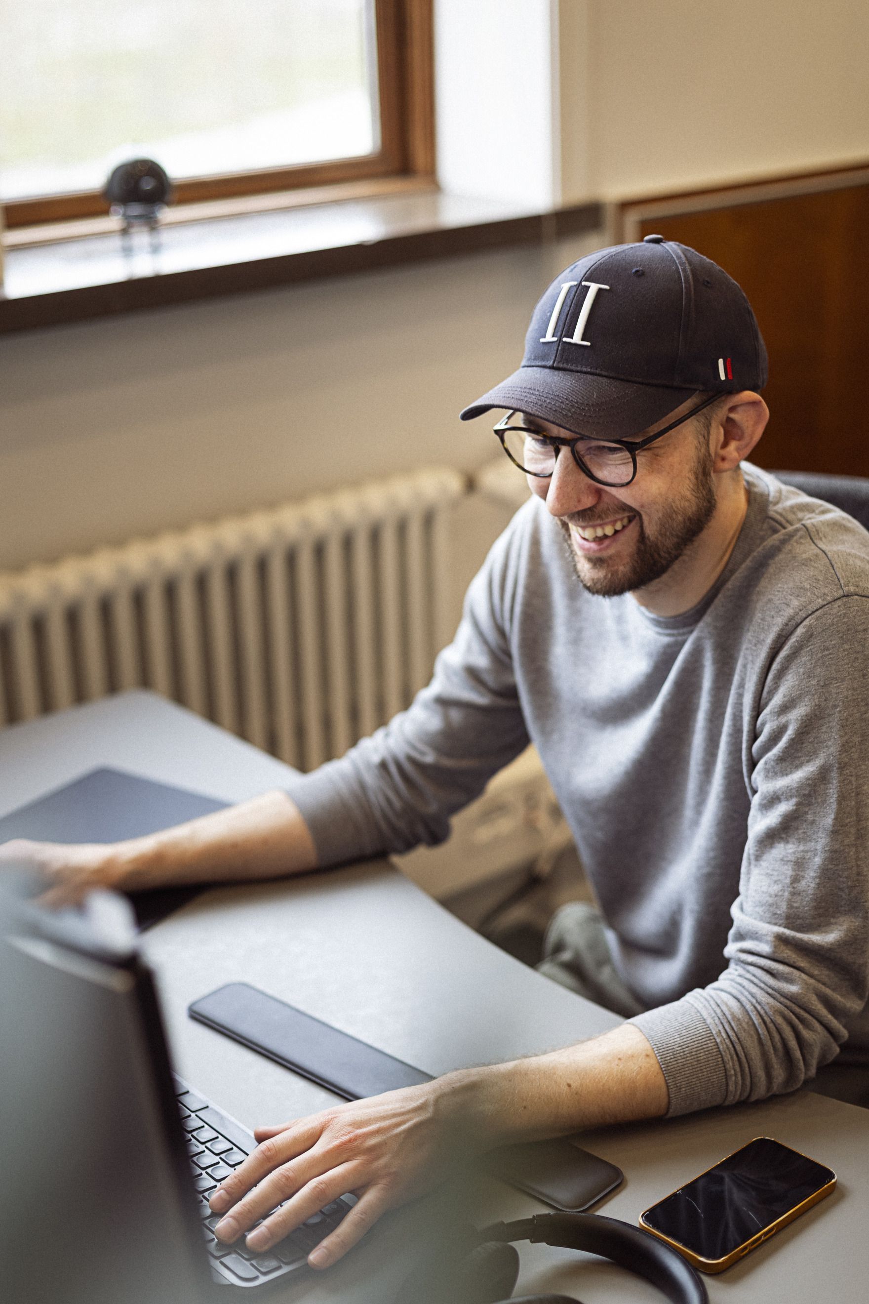 En mand med briller og kasket sidder foran en computer, smilende, mens han arbejder på en kontorarbejdsplads med en smartphone ved siden af.