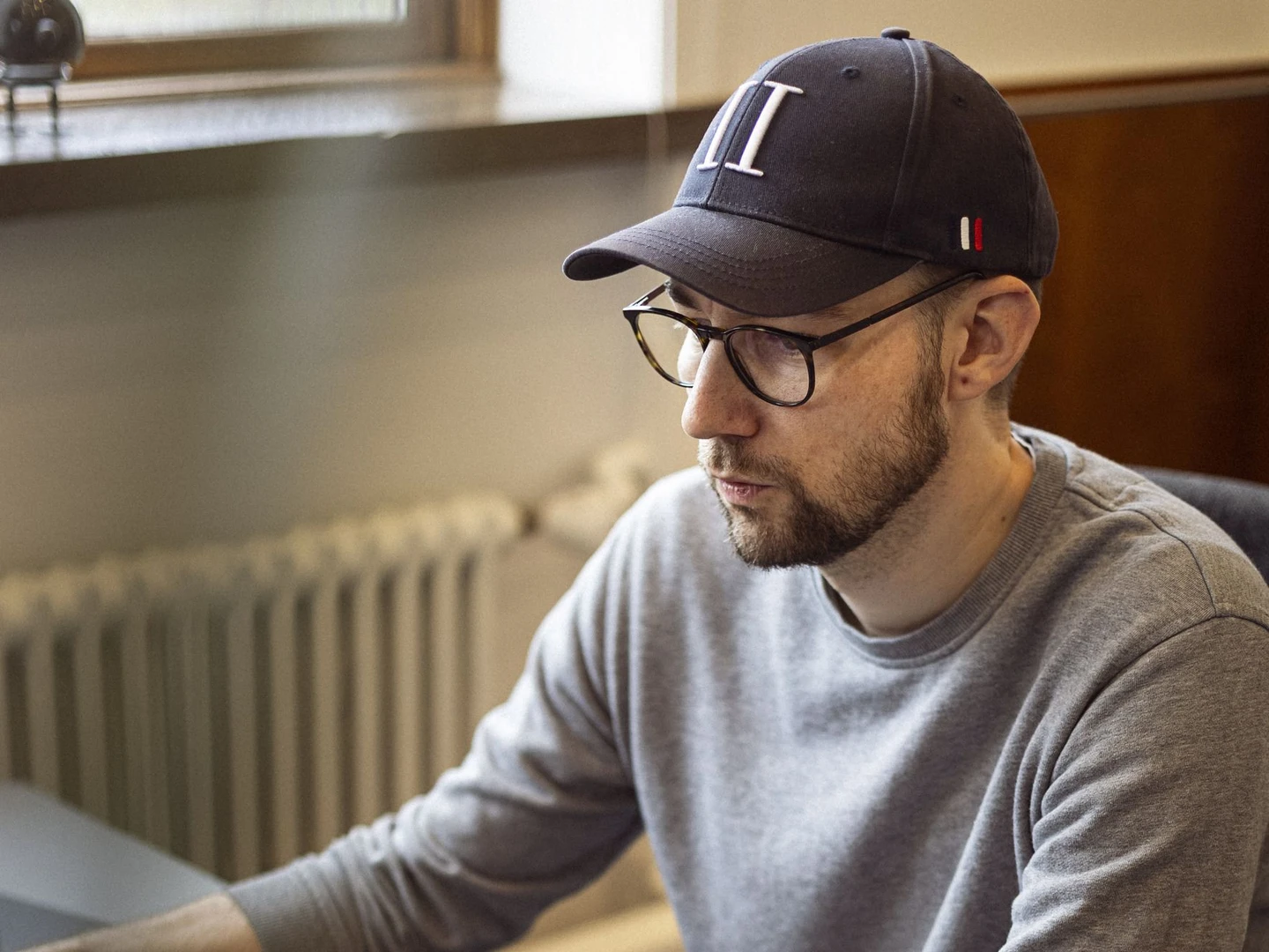 En mand sidder ved et skrivebord og arbejder på en bærbar computer, iført en grå sweater og en baseballkasket, med en mobiltelefon og høretelefoner på bordet.