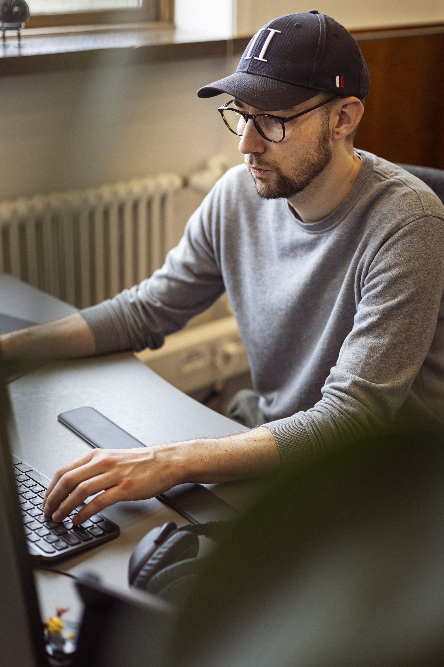 En mand sidder ved et skrivebord og arbejder på en bærbar computer, iført en grå sweater og en baseballkasket, med en mobiltelefon og høretelefoner på bordet.