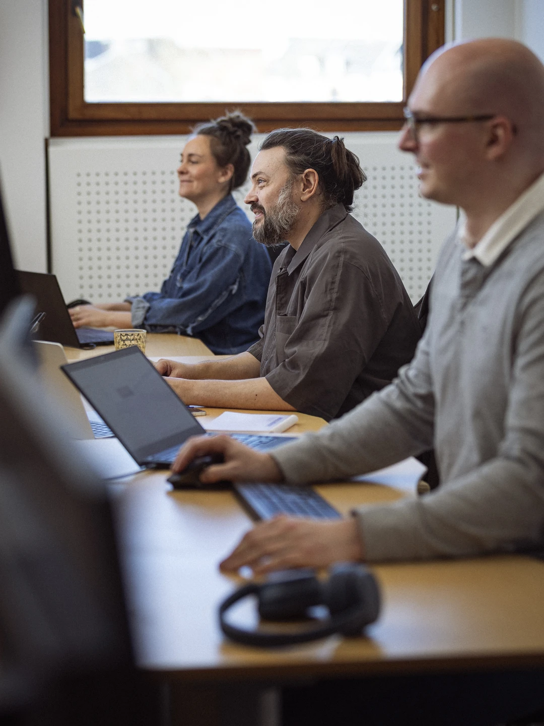 Tre personer arbejder koncentreret ved skriveborde med laptops og tilbehør, i et lyst kontormiljø med vinduer.
