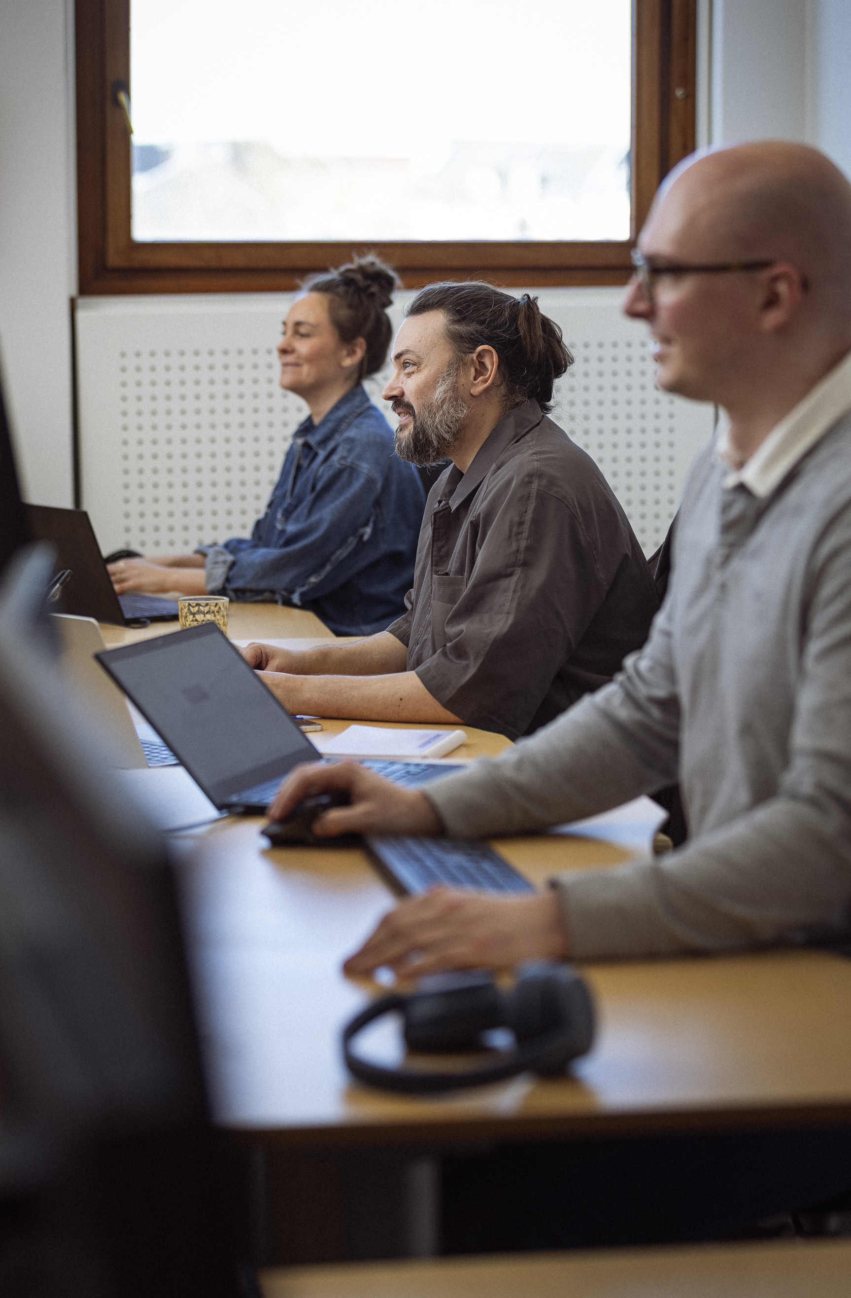 Tre personer arbejder koncentreret ved skriveborde med laptops og tilbehør, i et lyst kontormiljø med vinduer.
