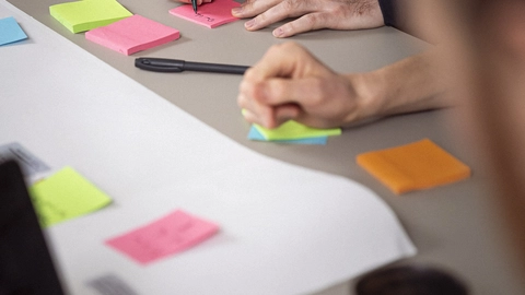 Two people engaged in collaborative brainstorming at a table covered in colorful sticky notes and a large sheet of paper, with one individual writing on a pink note and the other using a pen on a green note. A laptop and a small container are also visible in the foreground.