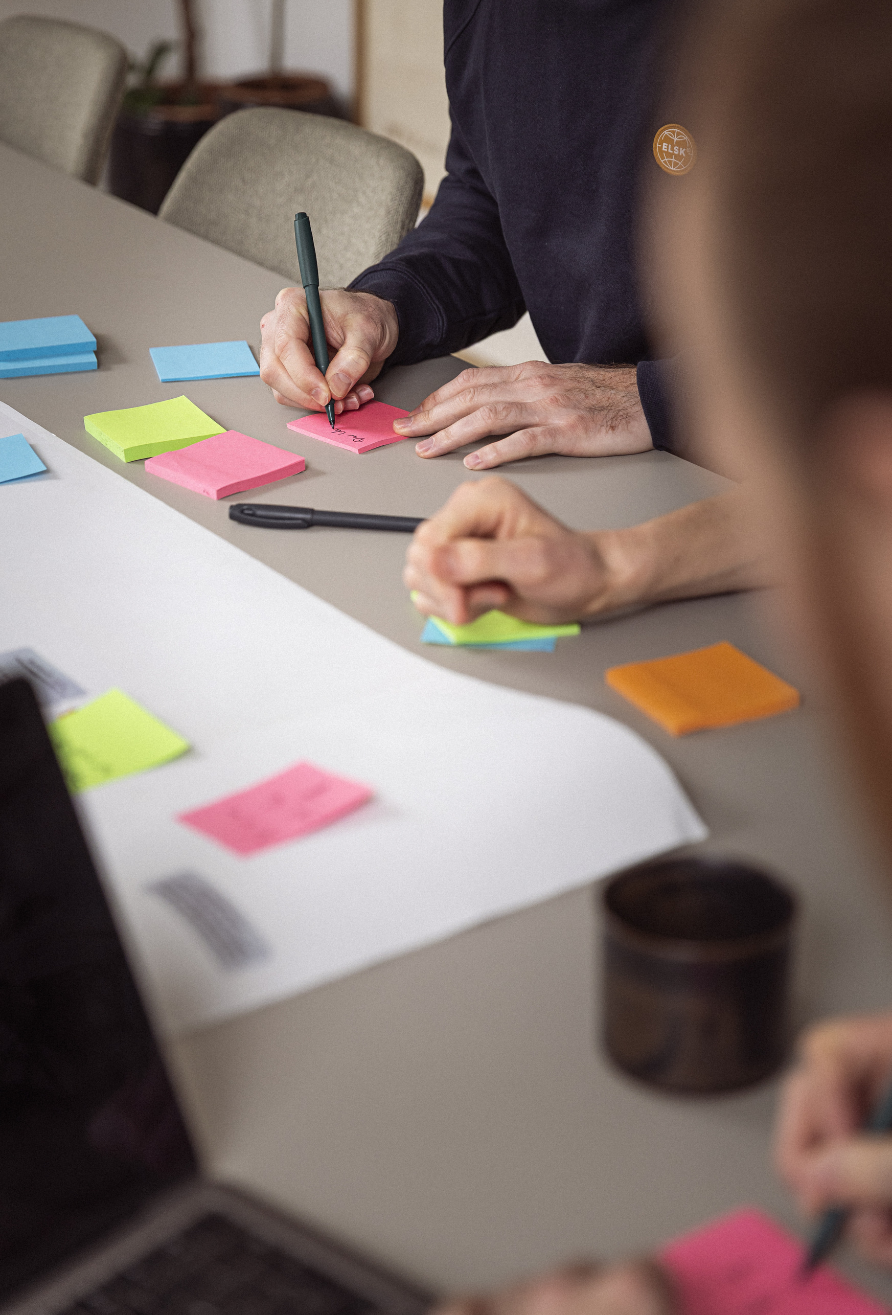 Hænder skriver på farvede post-it sedler under en brainstorming-session, med en bærbar computer og papir på bordet.