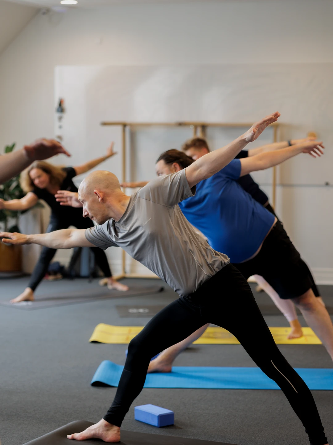 En gruppe mennesker deltager i en yogaklasse, hvor de udfører forskellige yogaøvelser på måtter i et lyst rum. Fokuseret mand i gråt træningstøj strækker sig i en krigerstilling, mens andre deltager ligeledes praktiserer yoga i baggrunden.