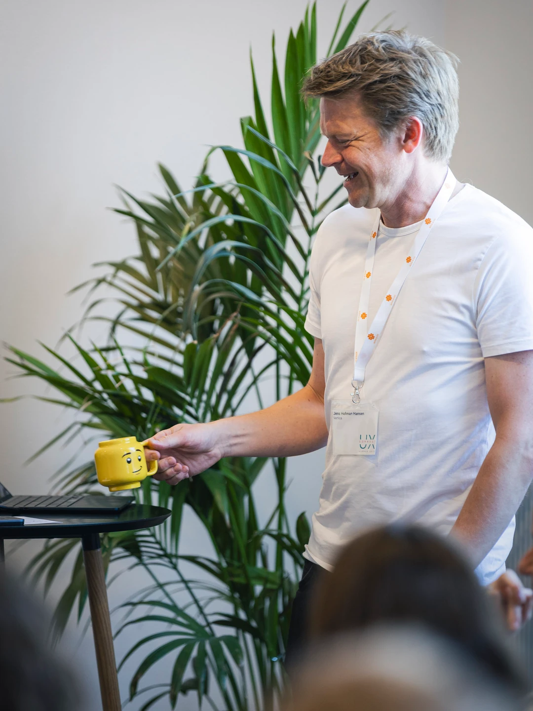 En smilende mand i hvid T-shirt holder en gul kop med et ansigtstræk, stående ved en laptop med grønne planter i baggrunden.