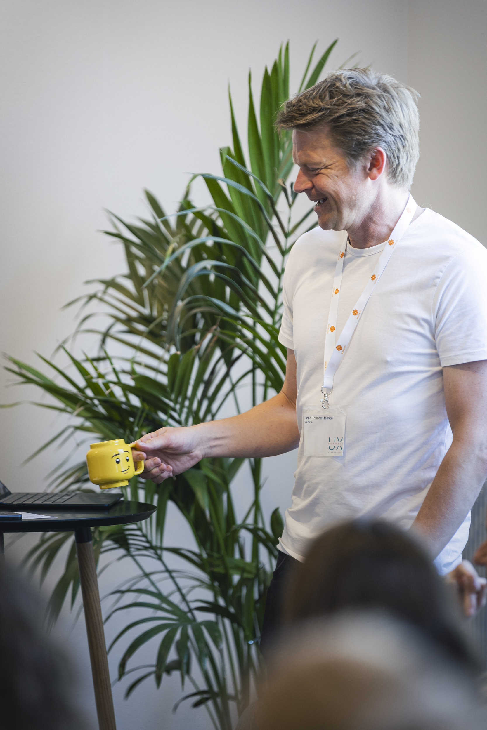 En smilende mand i hvid T-shirt holder en gul kop med et ansigtstræk, stående ved en laptop med grønne planter i baggrunden.