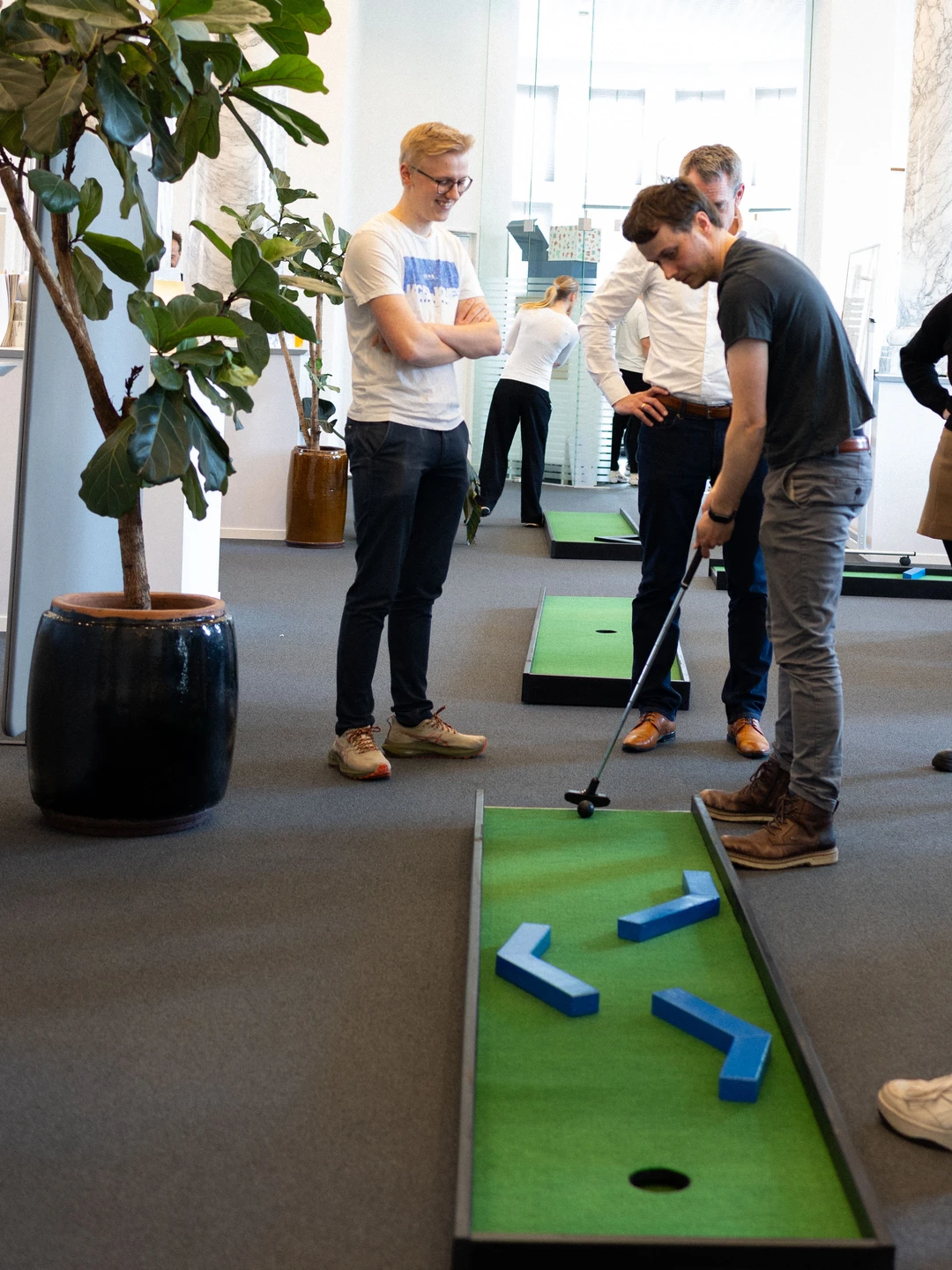 Mænd der spiller minigolf i et kontormiljø, omgivet af grønne planter og indretning, med fokus på en spiller der sigter mod en hul i banen.