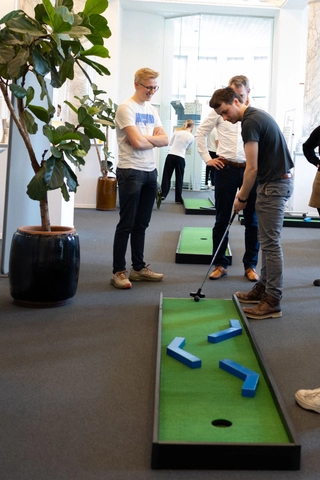 Mænd der spiller minigolf i et kontormiljø, omgivet af grønne planter og indretning, med fokus på en spiller der sigter mod en hul i banen.