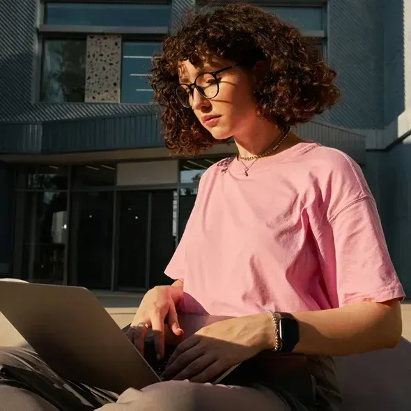 Ung kvinde med krøllede hår iført en lyserød t-shirt sidder koncentreret med en laptop, mens hun arbejder udendørs i et moderne bymiljø.