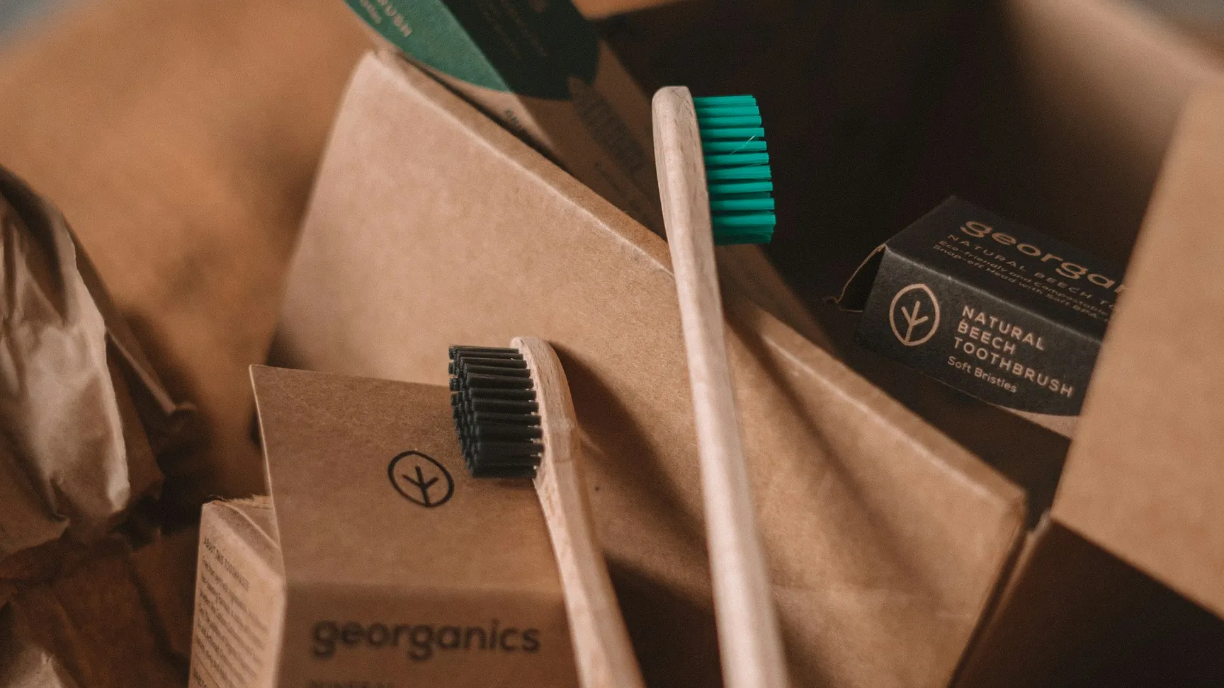 Billede af en naturlig tandbørste og tandpasta fra Georganics, præsenteret i en genanvendelig papkasse omgivet af brunt papir ved siden af en glasbeholder.