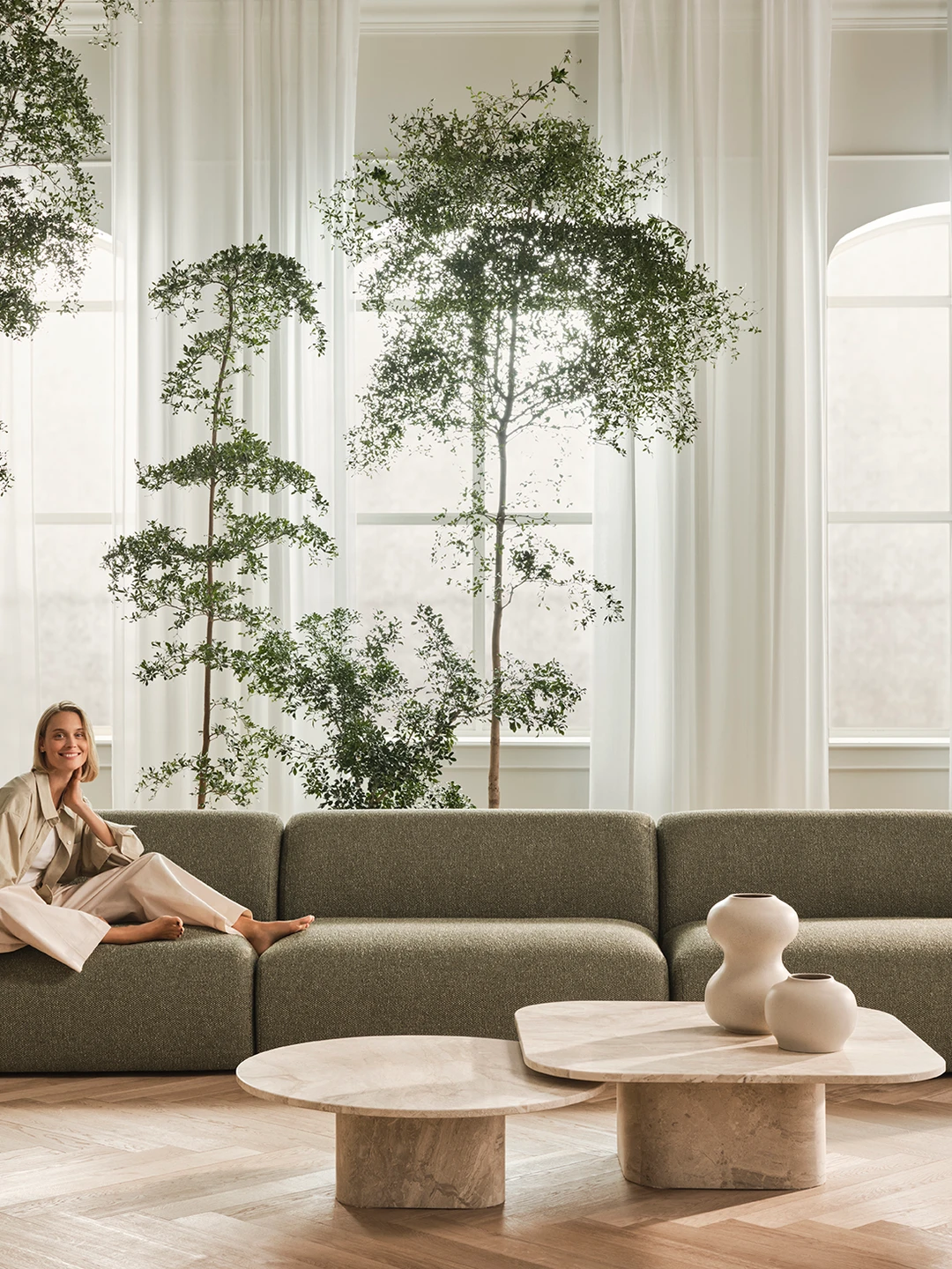A stylish woman sits comfortably on a modern green sofa in a bright, airy interior filled with indoor trees, large windows draped with sheer curtains, and minimalistic decorative elements including round coffee tables and elegant planters.
