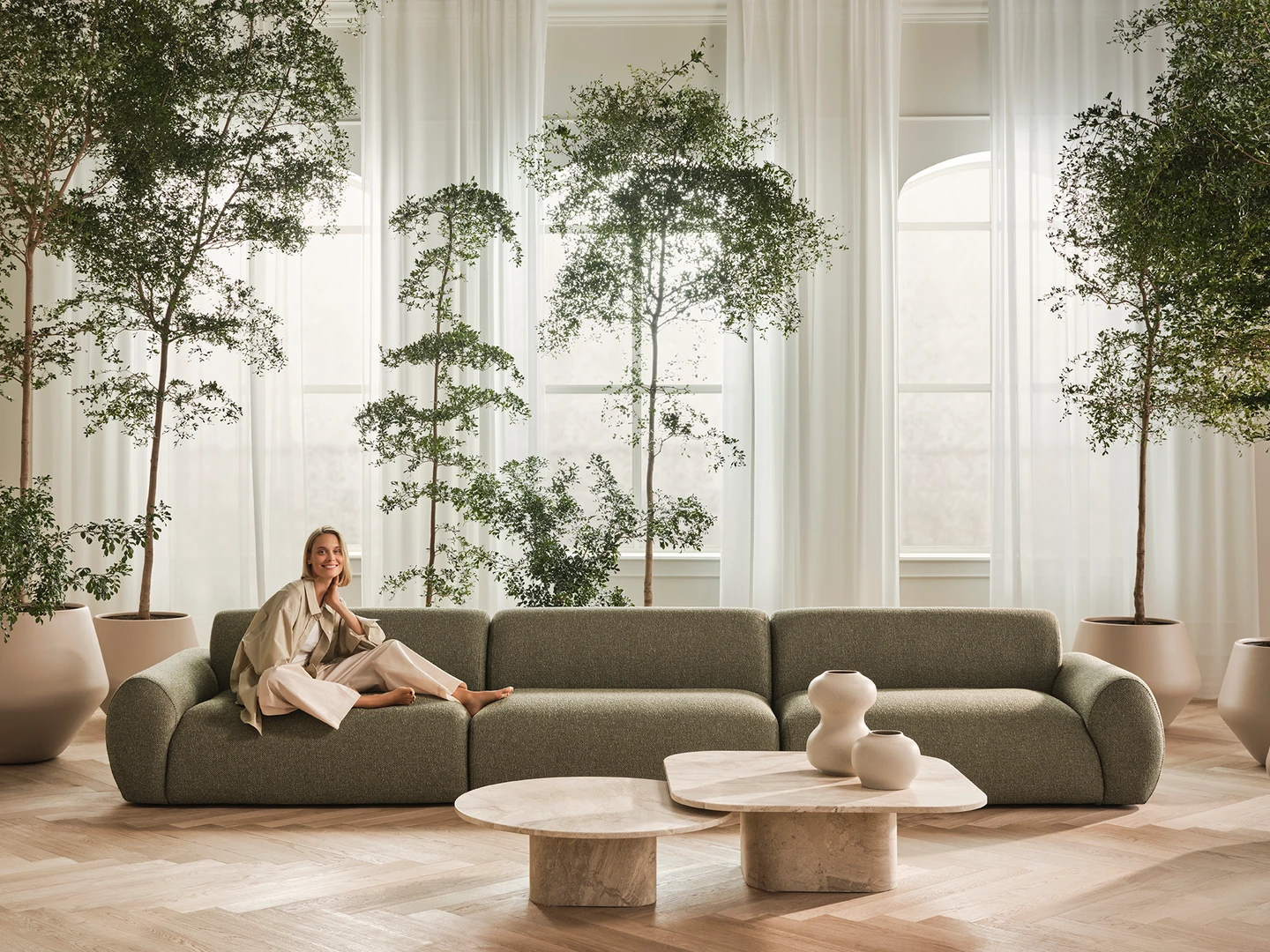 En moderne stue med en grøn sofa, en kvinde iført lyse tøj der smiler, store planter i potter, og et elegant marmorkaffebord.