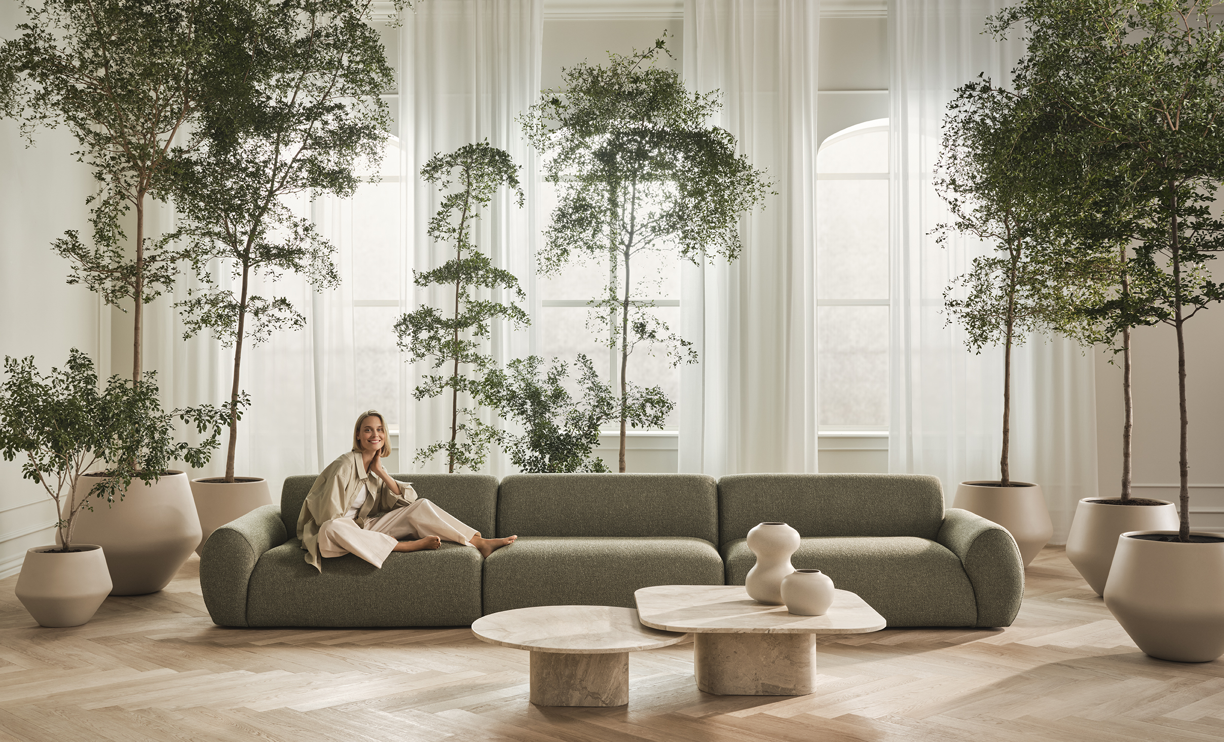 En moderne stue med en grøn sofa, en kvinde iført lyse tøj der smiler, store planter i potter, og et elegant marmorkaffebord.