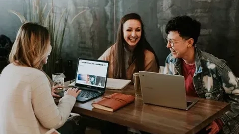Tre unge kvinder sidder ved et bord med bærbare computere, smilende og griner, med noter og en vandglas synligt; en afslappet atmosfære i et moderne rum.