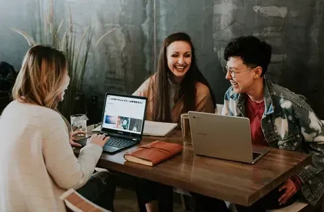 Tre unge kvinder sidder ved et bord med bærbare computere, smilende og griner, med noter og en vandglas synligt; en afslappet atmosfære i et moderne rum.