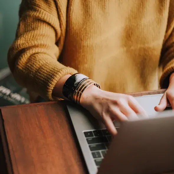 En hånd arbejder på en laptop, mens en person iført en gul sweater sidder ved et bord. Billede viser også smykker på håndleddet, der tilføjer et stilfuldt element til den afslappede arbejdsindstilling.