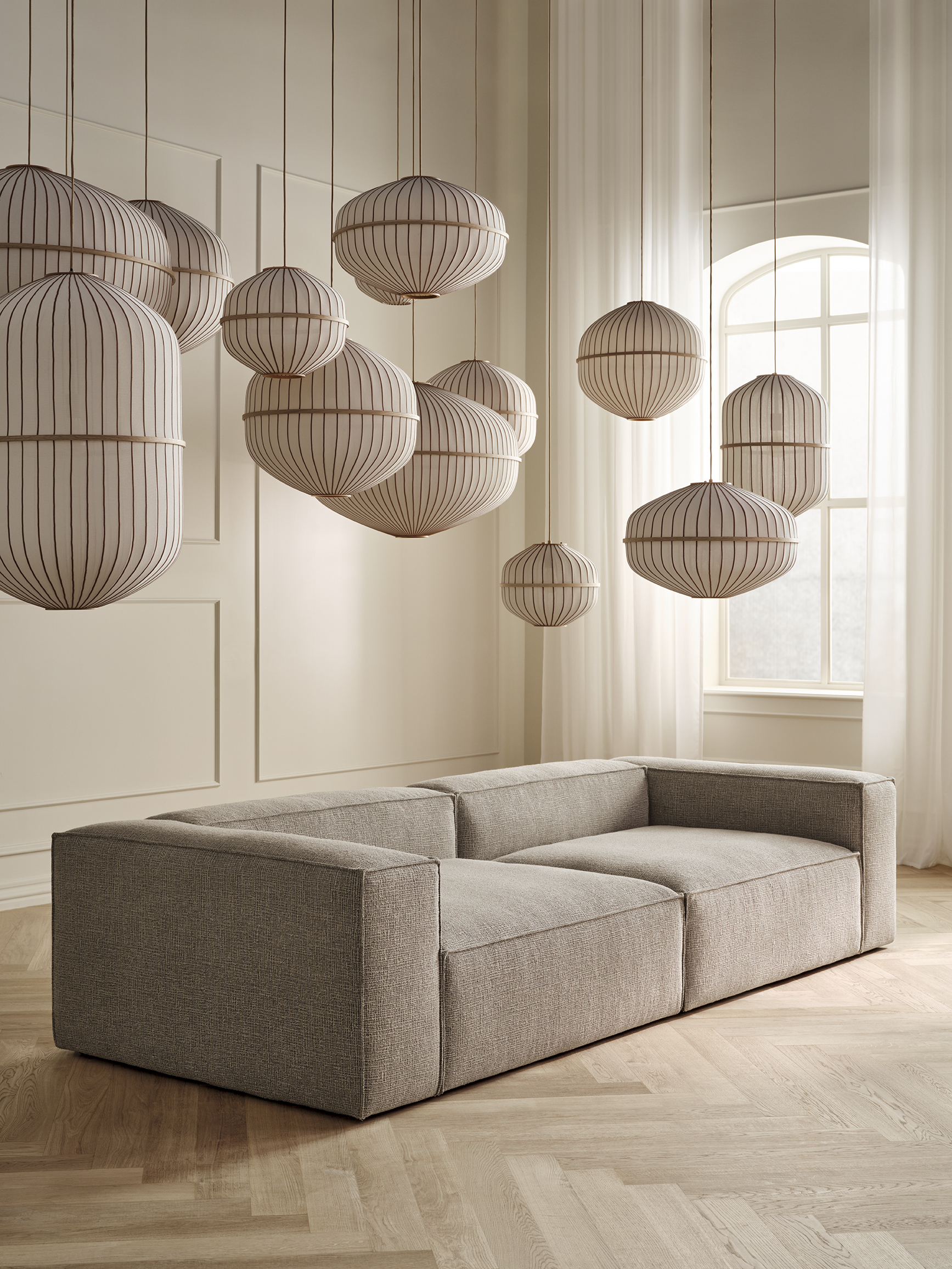 Sofasæt i minimalistisk stil med grå polstring, belejligt placeret under moderne lampedesign i et lyst rum med store vinduer og strømlinede vægge.