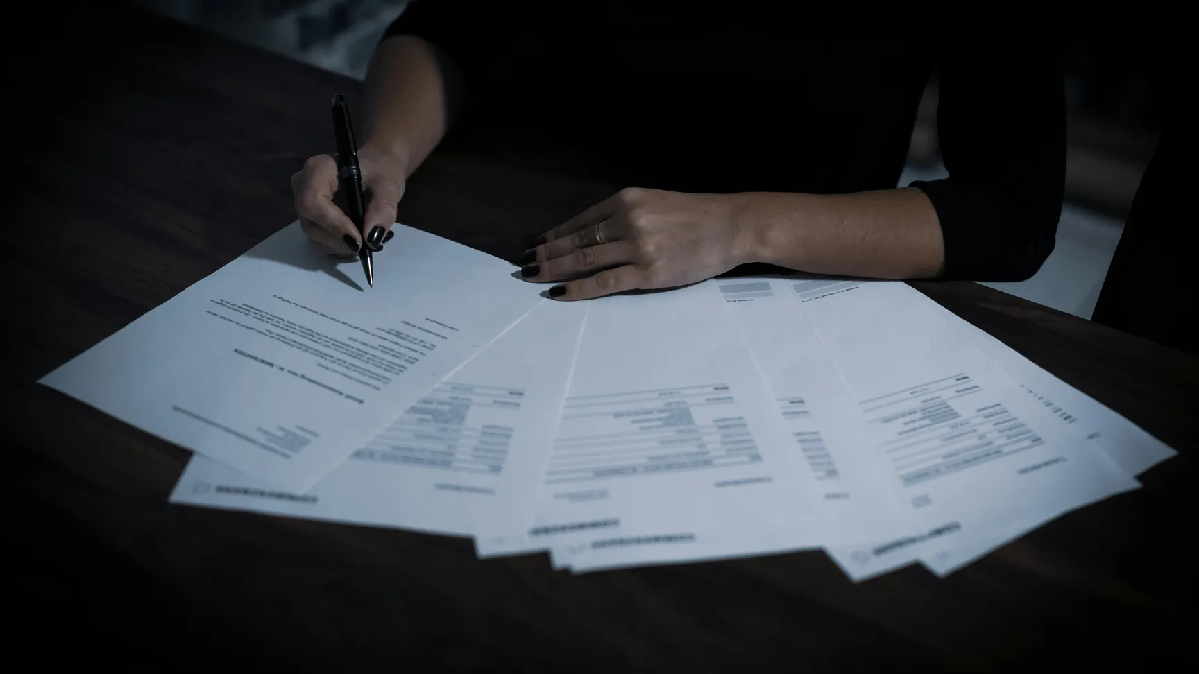 En person skriver med en sort pen på et dokument, mens der ligger flere papirer på et træbord, der indeholder oplysninger og regninger.