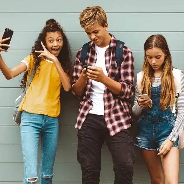 Tre unge mennesker står op ad en væg; en pige tager en selfie med en smartphone, mens en dreng i midten ser på sin mobiltelefon, og en anden pige til højre tjekker sin telefon.
