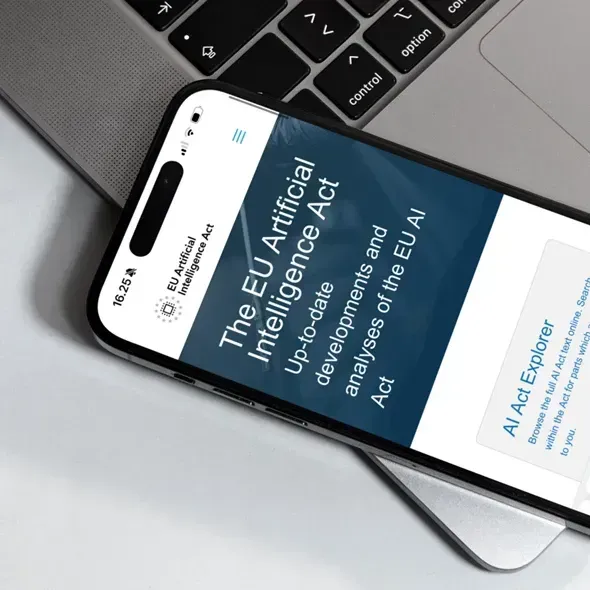 En smartphone viser hjemmesiden for EU's lovgivning om kunstig intelligens med opdateringer og analyser af loven, placeret på et skrivebord ved siden af en bærbar computer.