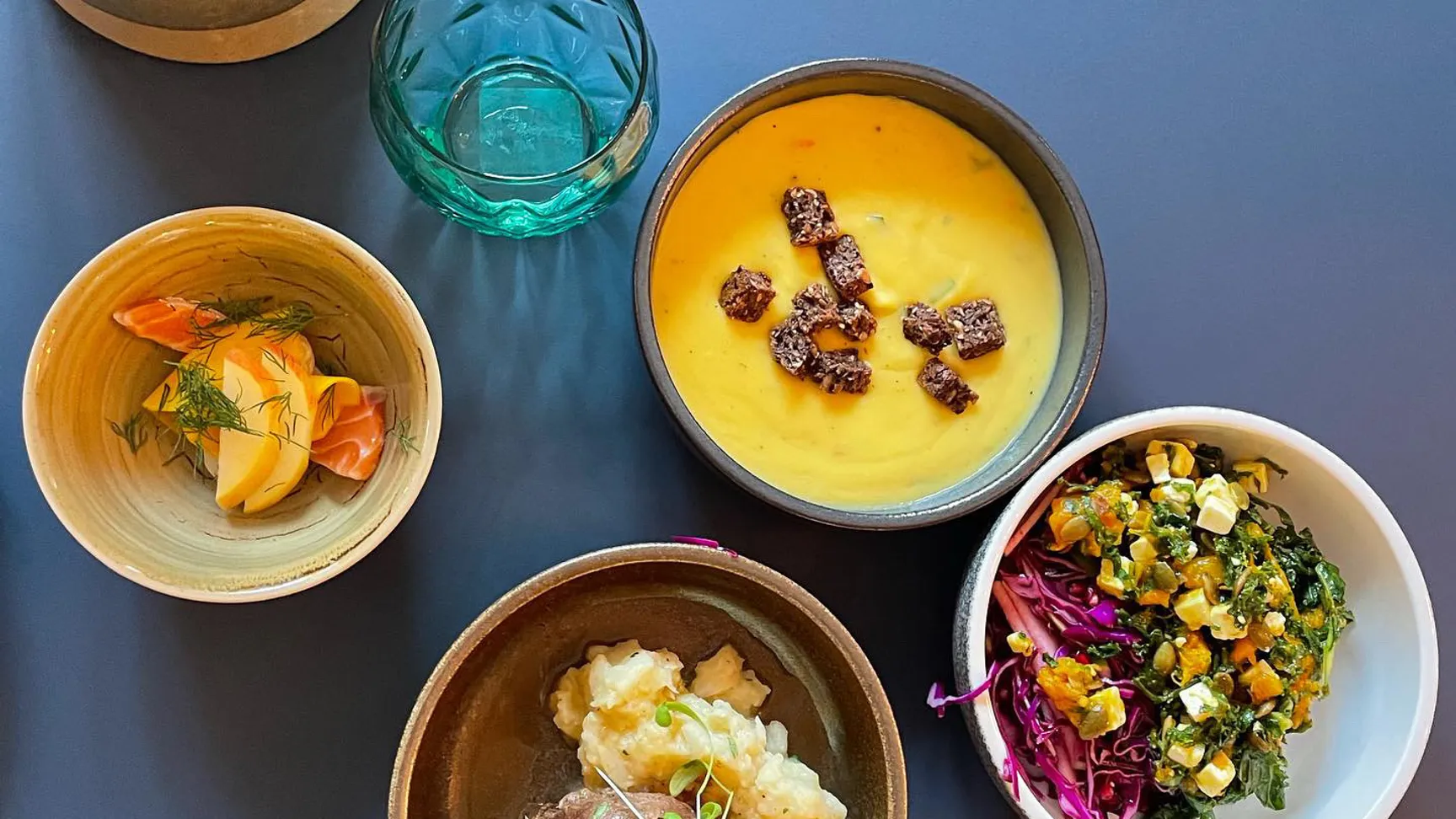 En farverig tallerken med flere retter, herunder en gul suppe med toppings, en portion kartoffelmos med kød, en skål med blandede grøntsager og en sideanretning med frugt og urter, præsenteret på en blå bordplade sammen med glas og karaffel.