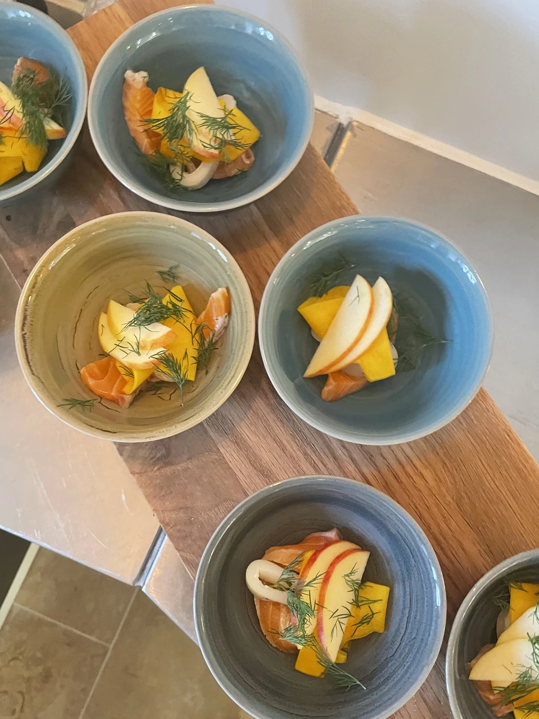 Skåle med friske æbler og mango samt røget laks, pyntet med dild, placeret på en træplade.
