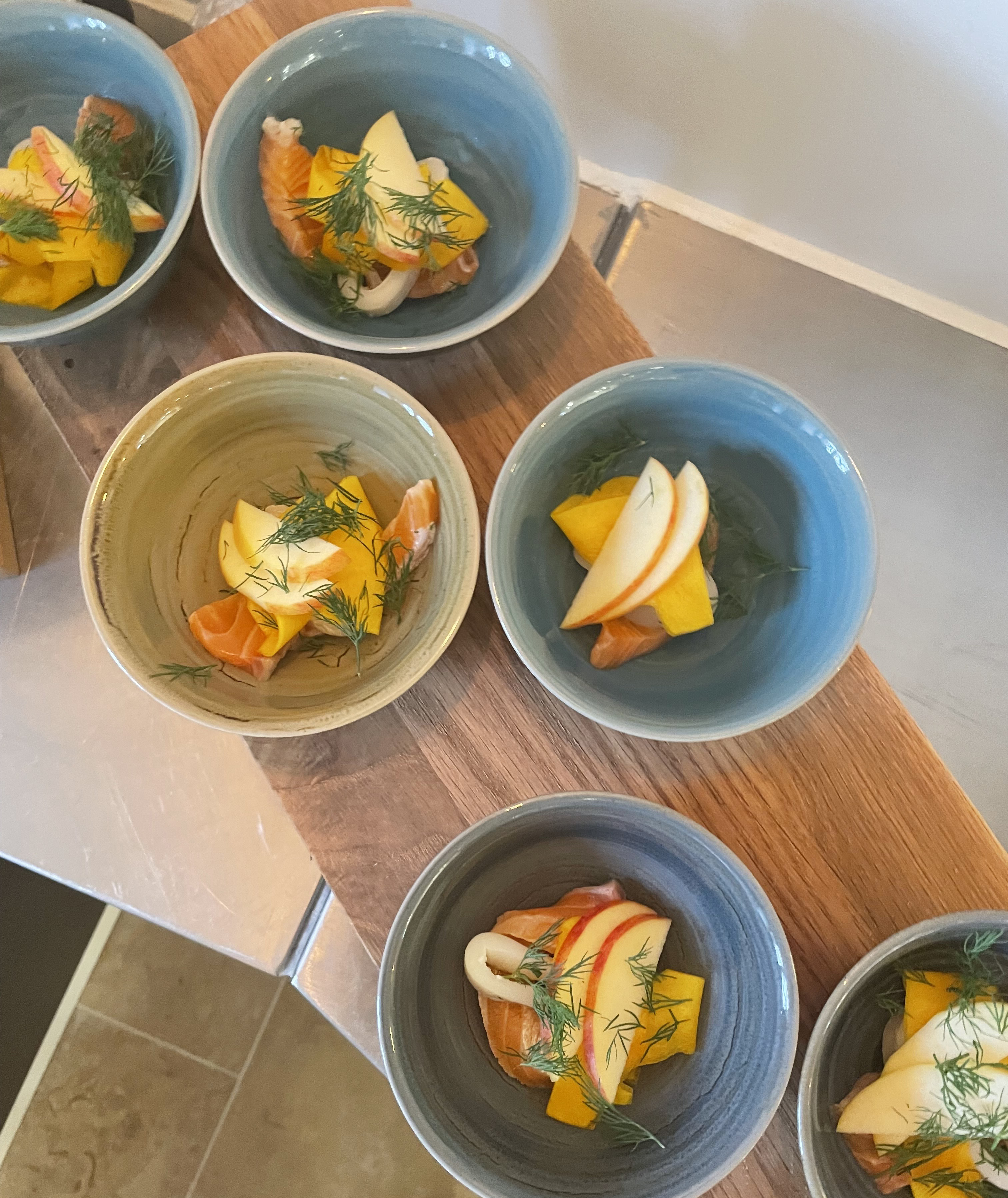 Skåle med friske æbler og mango samt røget laks, pyntet med dild, placeret på en træplade.