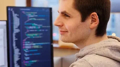 Ung mand med gråt hættesweater smiler, mens han arbejder ved computer med kode på skærmen.
