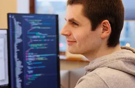 Ung mand med gråt hættesweater smiler, mens han arbejder ved computer med kode på skærmen.