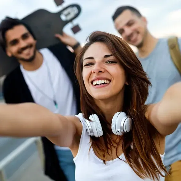 Unge mennesker tager et selfie udendørs med en skateboard i baggrunden, den kvindelige hovedperson har hovedtelefoner på og smiler bredt, mens to mænd står tættere på.